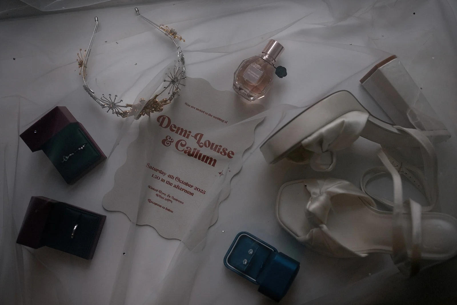 Wedding invitation card for Demi-Louise and Callum, a pair of white wedding shoes, a tiara, a bottle of perfume, two open ring boxes, a pair of white slippers, and a container of makeup brushes arranged on a white fabric surface.
