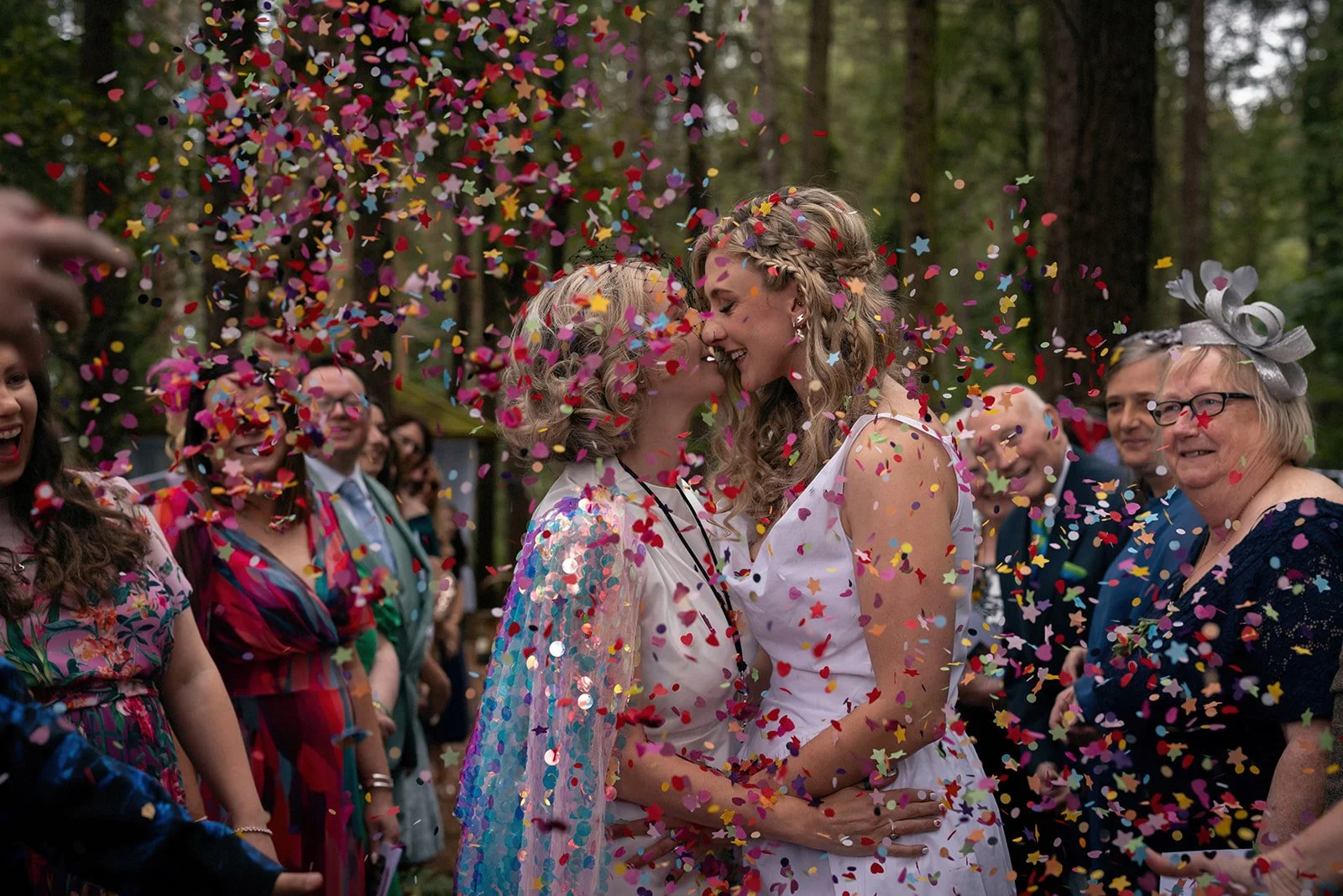 Two women, with their foreheads touching, are celebrating at an outdoor wedding surrounded by guests. They are smiling amid colorful confetti falling in the air. The scene is set in a wooded area with trees in the background.