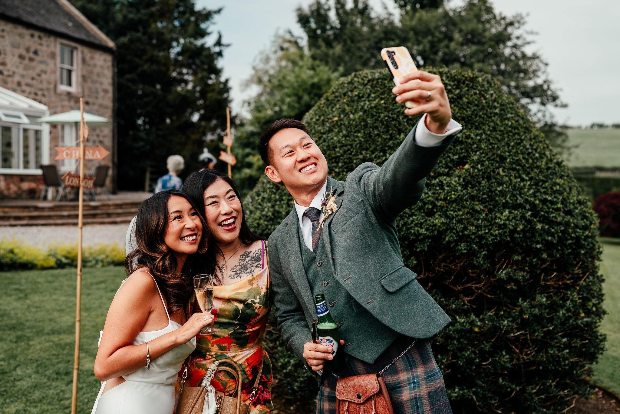 Three diverse friends in formal attire smiling and taking a selfie outdoors at a celebration, with a stone house and a green bush in the background.