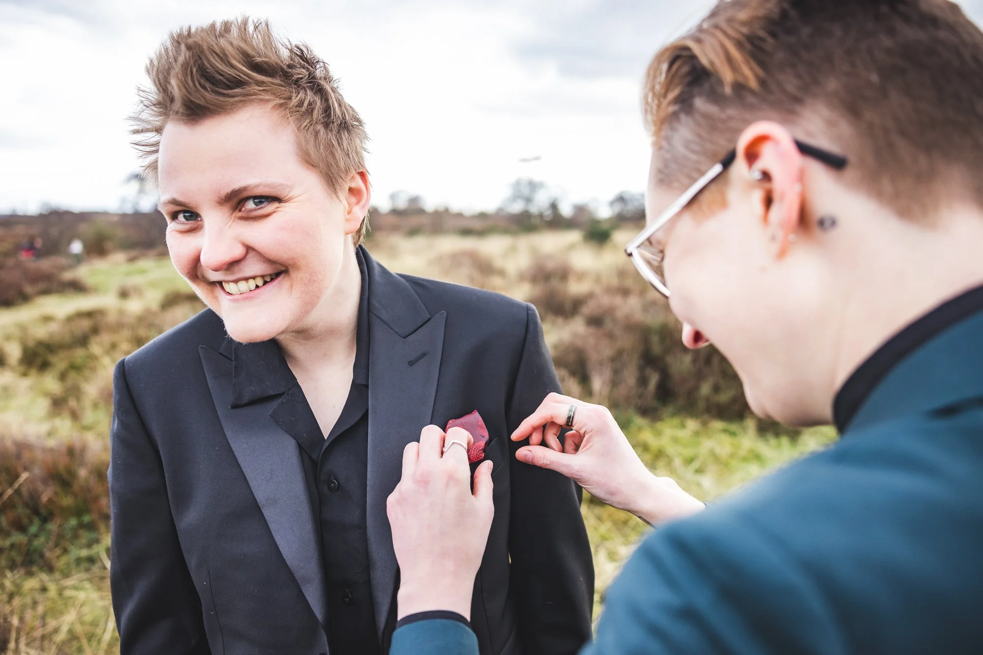Two women outdoors, one with short hair smiling while the other, with glasses and earrings, pins a red flower to her black blazer.