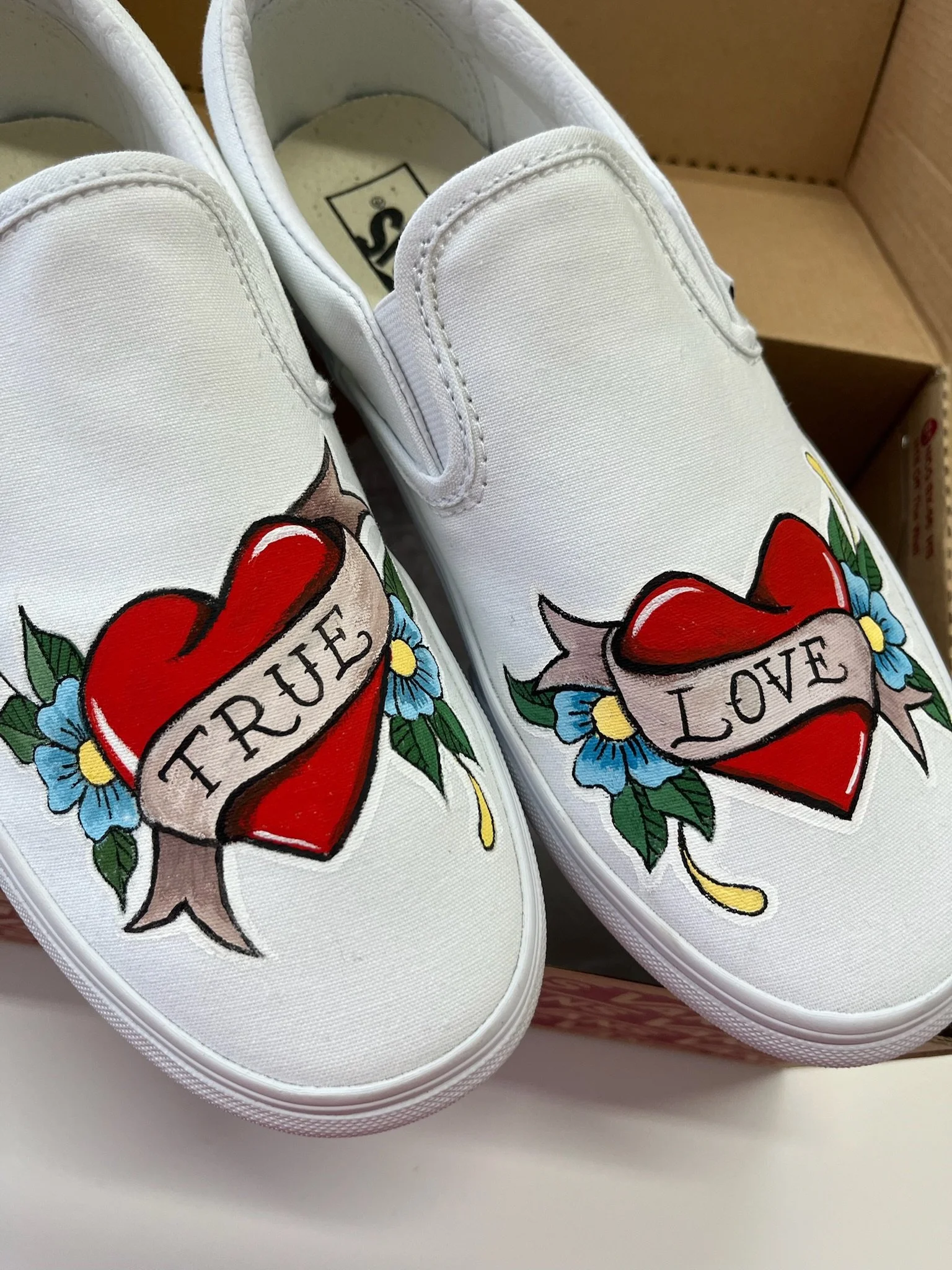White sneakers with tattoo-style tattoos of red hearts with banners reading "True" and "Love" and blue flowers.