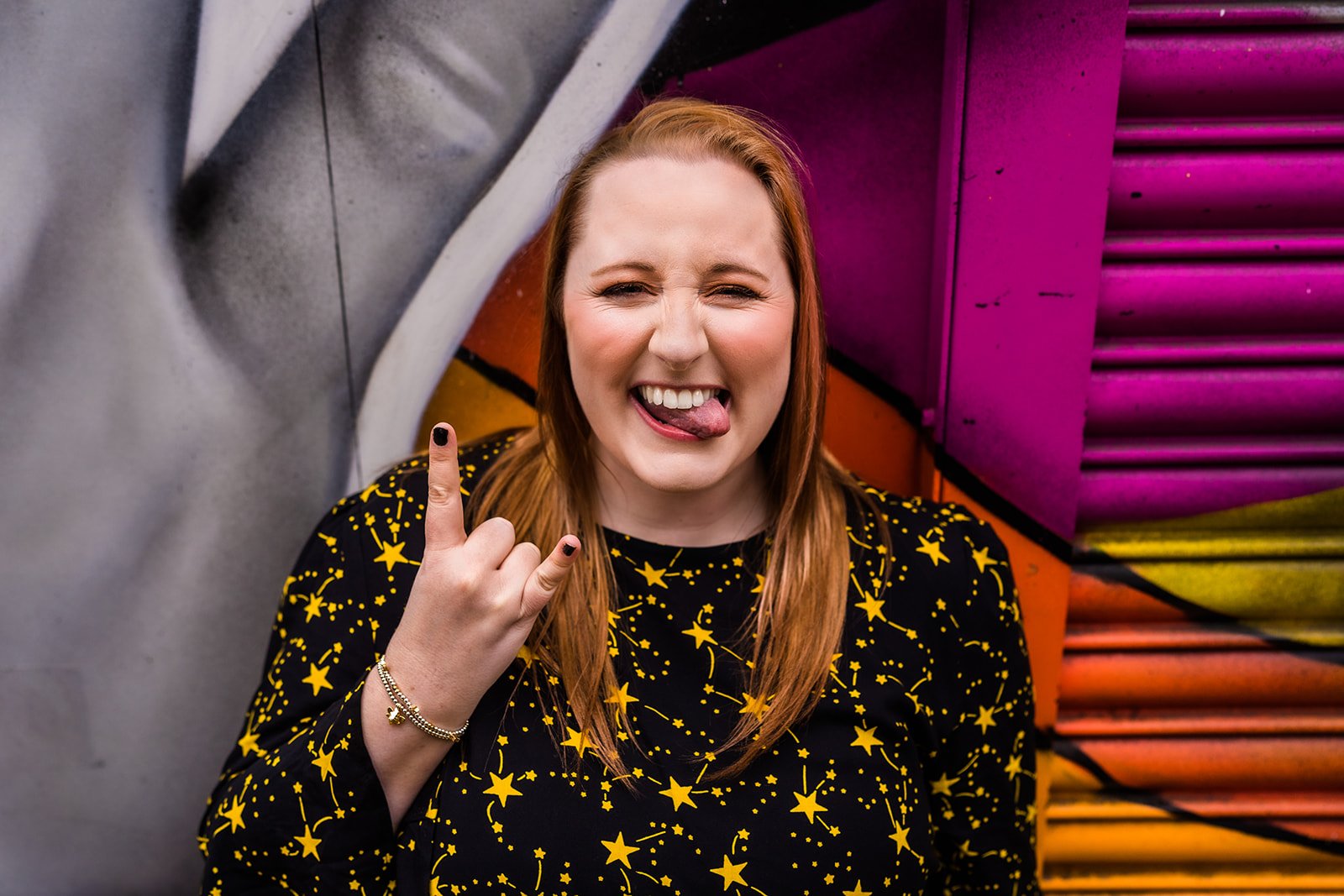 A young woman with red hair making a rock on hand gesture, sticking out her tongue, in front of colorful graffiti art.