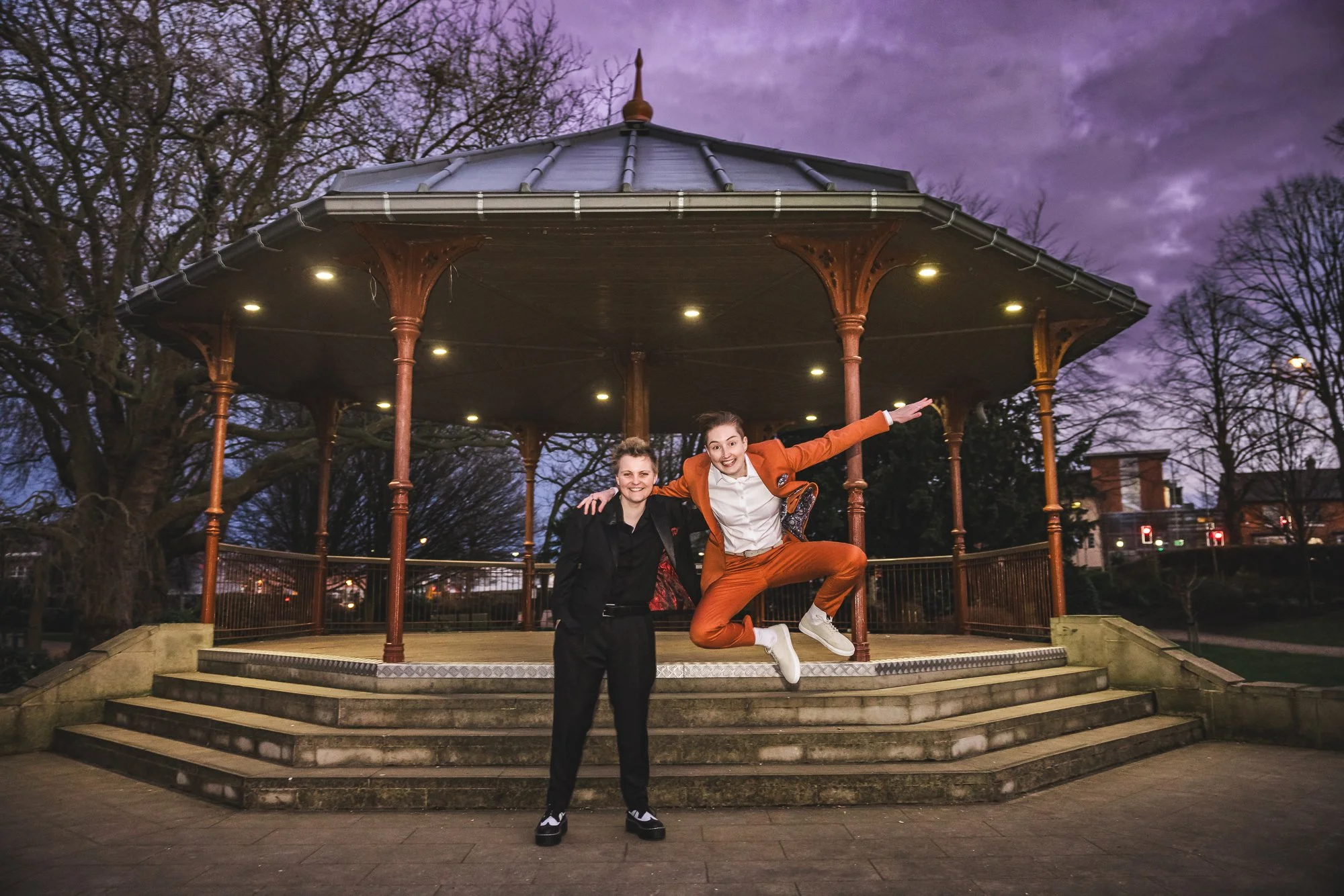Two women are in front of an ornate gazebo in a park during twilight. One woman is standing with an arm around the other's shoulder, while the other is mid-jump with arms outstretched. Both are smiling and dressed stylishly.