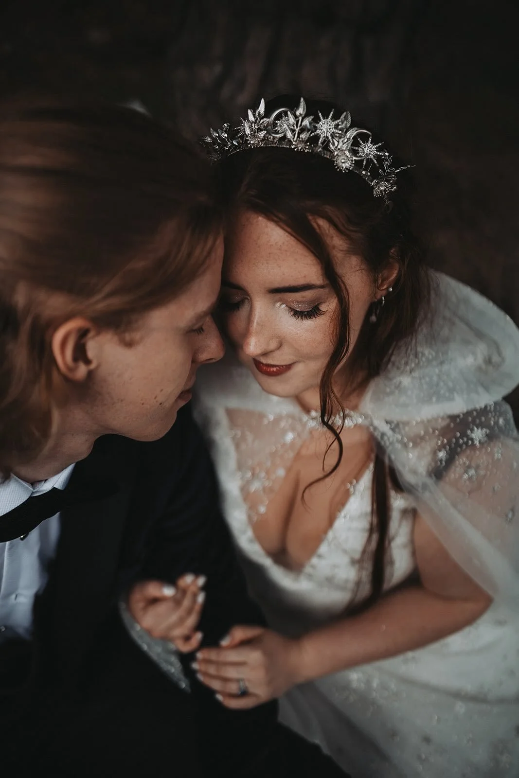 Close-up of a bride and groom with their foreheads touching, eyes closed, and smiling softly, dressed in wedding attire, with the bride wearing a tiara and the groom in a suit.