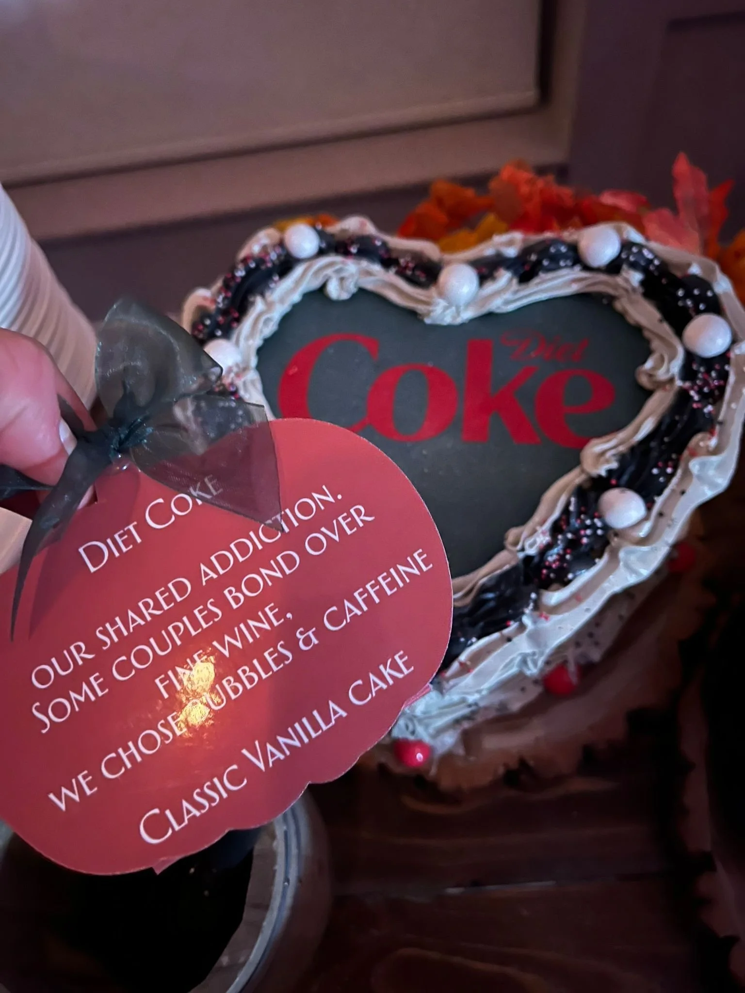 Heart-shaped cake with black, white, and red decorations, featuring the word "Coke" on top, and a black and red note in front says it is a vanilla cake shared over wine, bubbly, and caffeine.