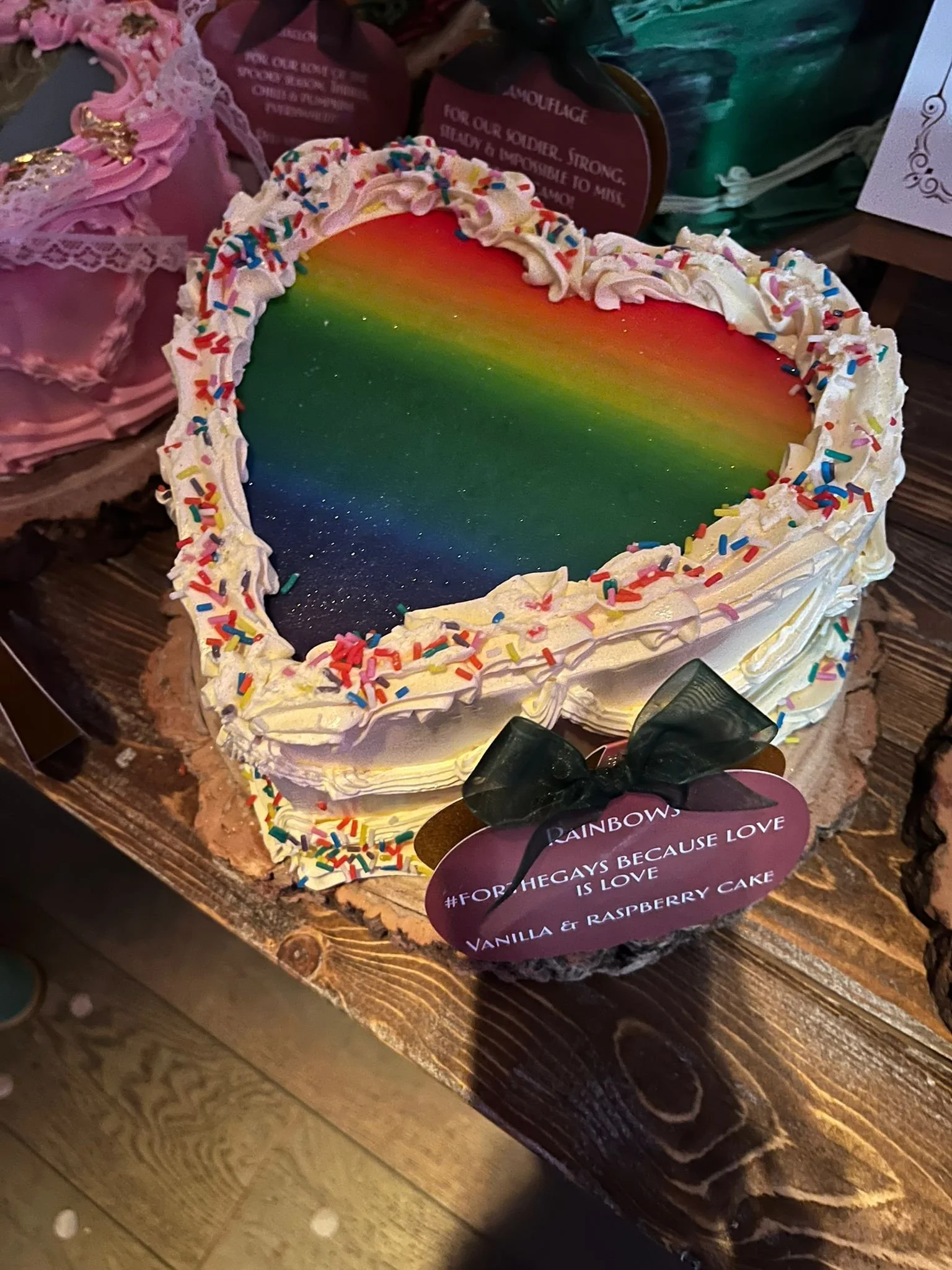 Colorful heart-shaped cake with rainbow design, white frosting, rainbow sprinkles, and a black ribbon with a purple heart-shaped tag that reads '#FOR THE GAYS BECAUSE LOVE IS LOVE Vanilla & Raspberry CAKE'.