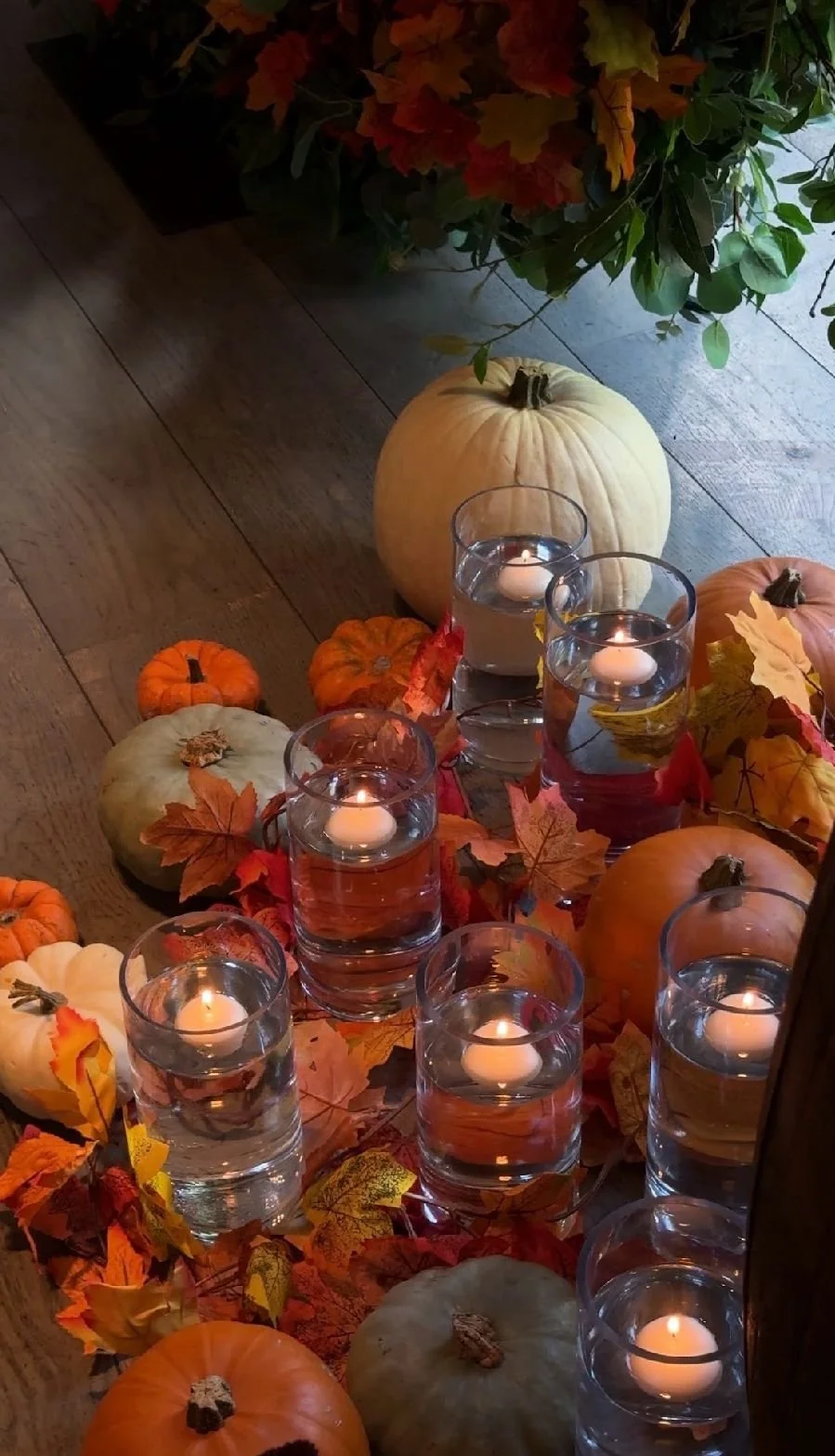 Autumn-themed display with pumpkins, glass candles, and fall leaves on a wooden surface.