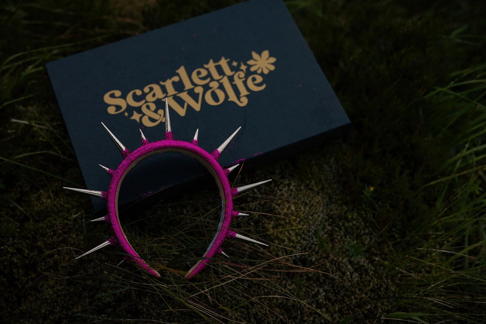 A black box with gold text saying 'Scarlett & Wolfie' and a flower decoration, lying on dark, mossy ground. A pink headband with white spikes is placed next to it.