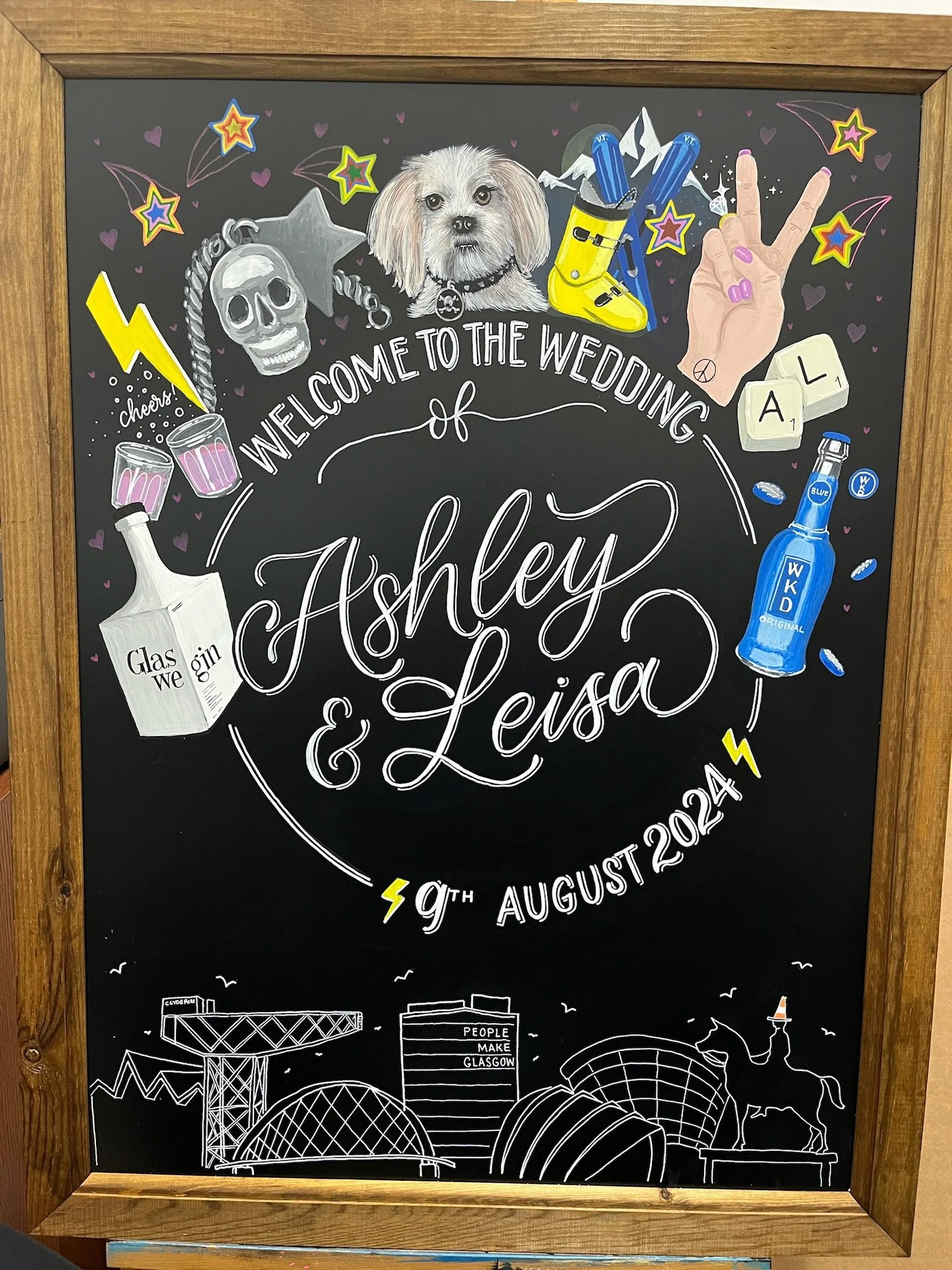 A decorative wedding announcement chalkboard featuring illustrations of a dog, skull, spray paint cans, bottles, a hand making a peace sign, a pair of dice, and symbols, with Edinburgh skyline at the bottom and the names Ashley and Leisa, and the date August 9th, 2024.