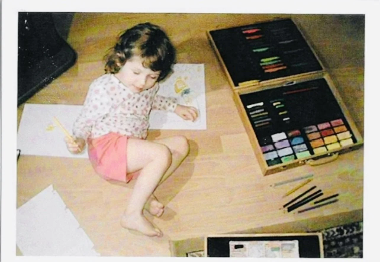 A young girl with curly brown hair drawing on a sheet of paper with colored pencils scattered around. A box of pastel chalks or oil pastels is open next to her, and there are additional sheets of paper nearby on a wooden floor.