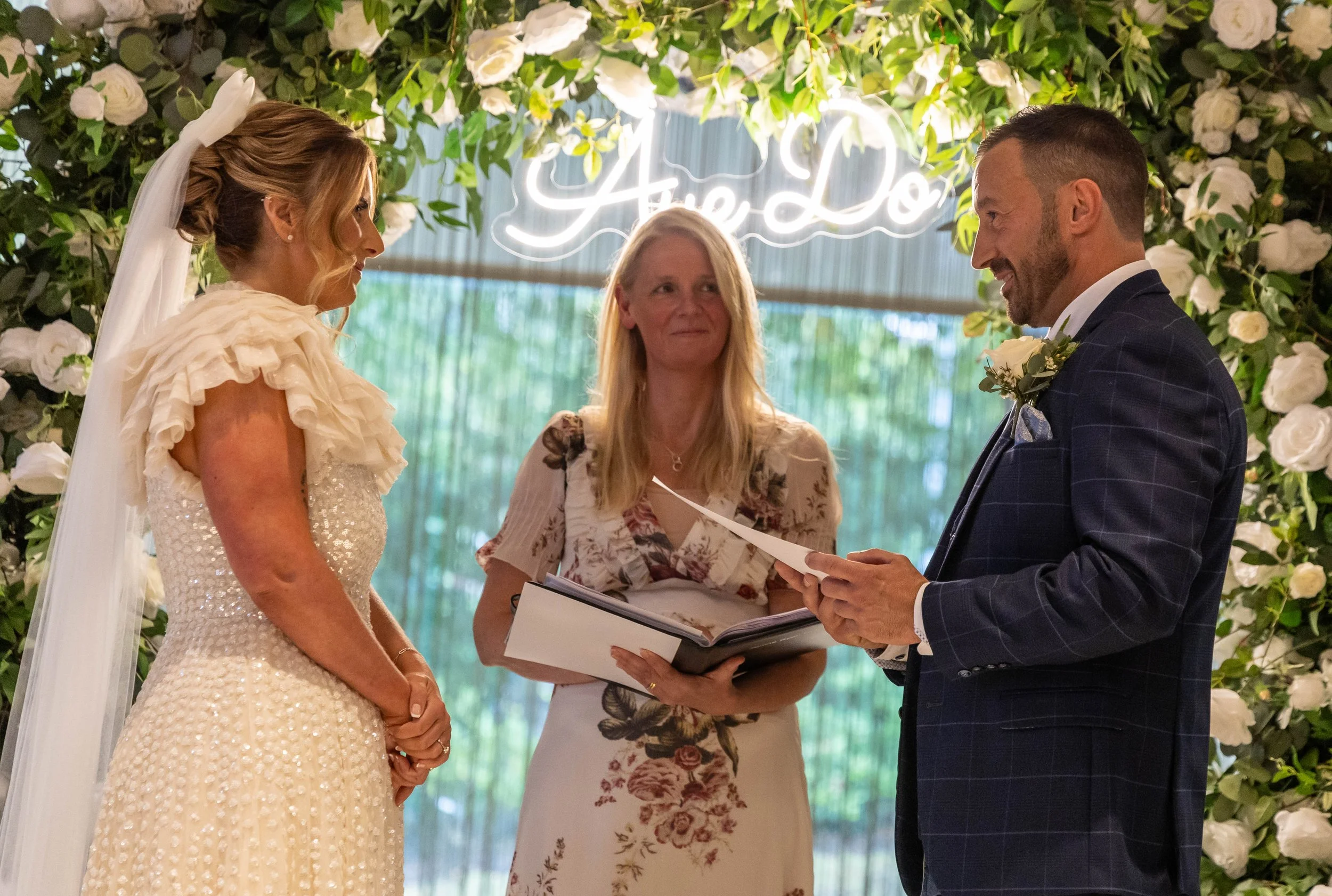 A bride and groom exchanging vows during a wedding ceremony with an officiant, surrounded by a floral backdrop with white roses and greenery, and a neon sign in the background.
