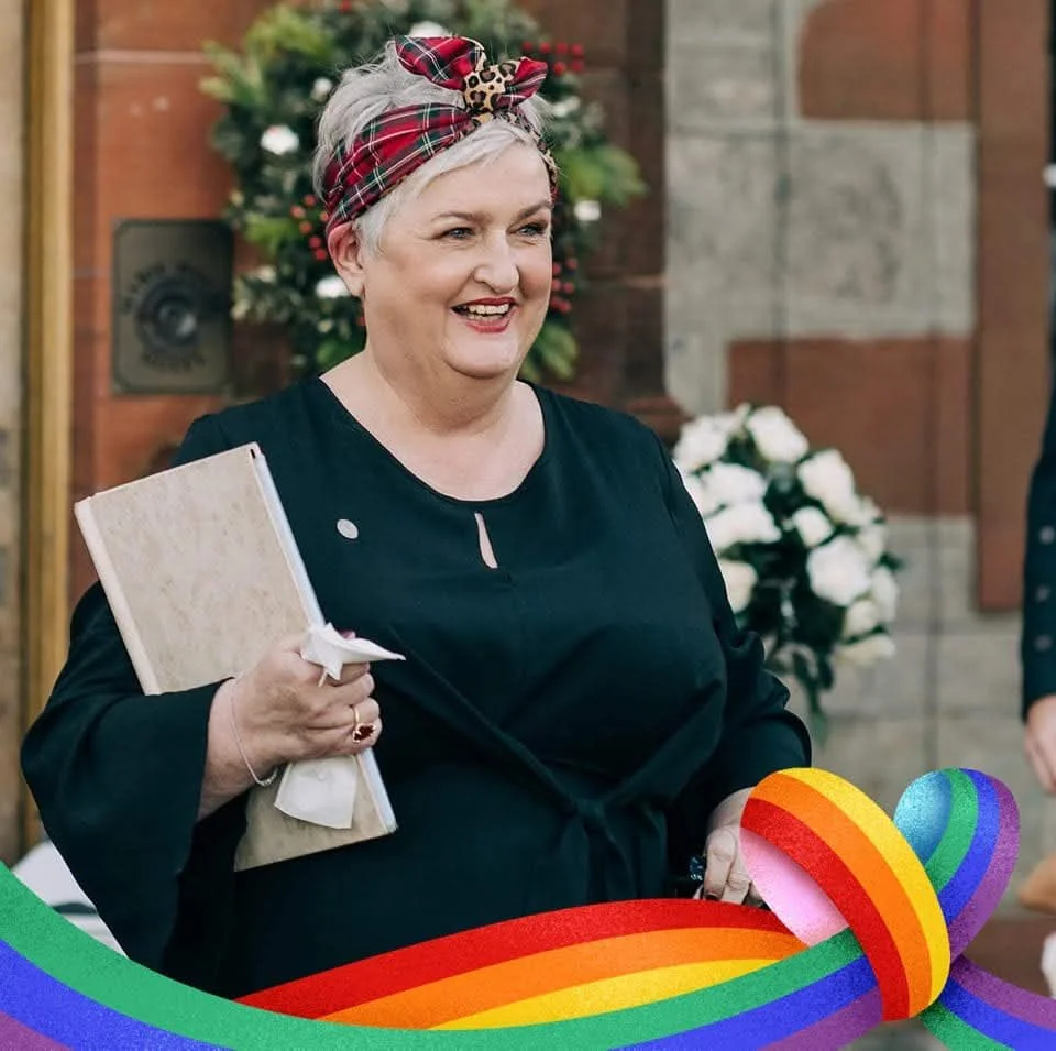 A smiling woman with short gray hair, wearing a plaid headband with leopard print accents, holding a menu or folder, standing in front of a decorated holiday wreath with white flowers. There is a rainbow-colored ribbon at the bottom of the image.