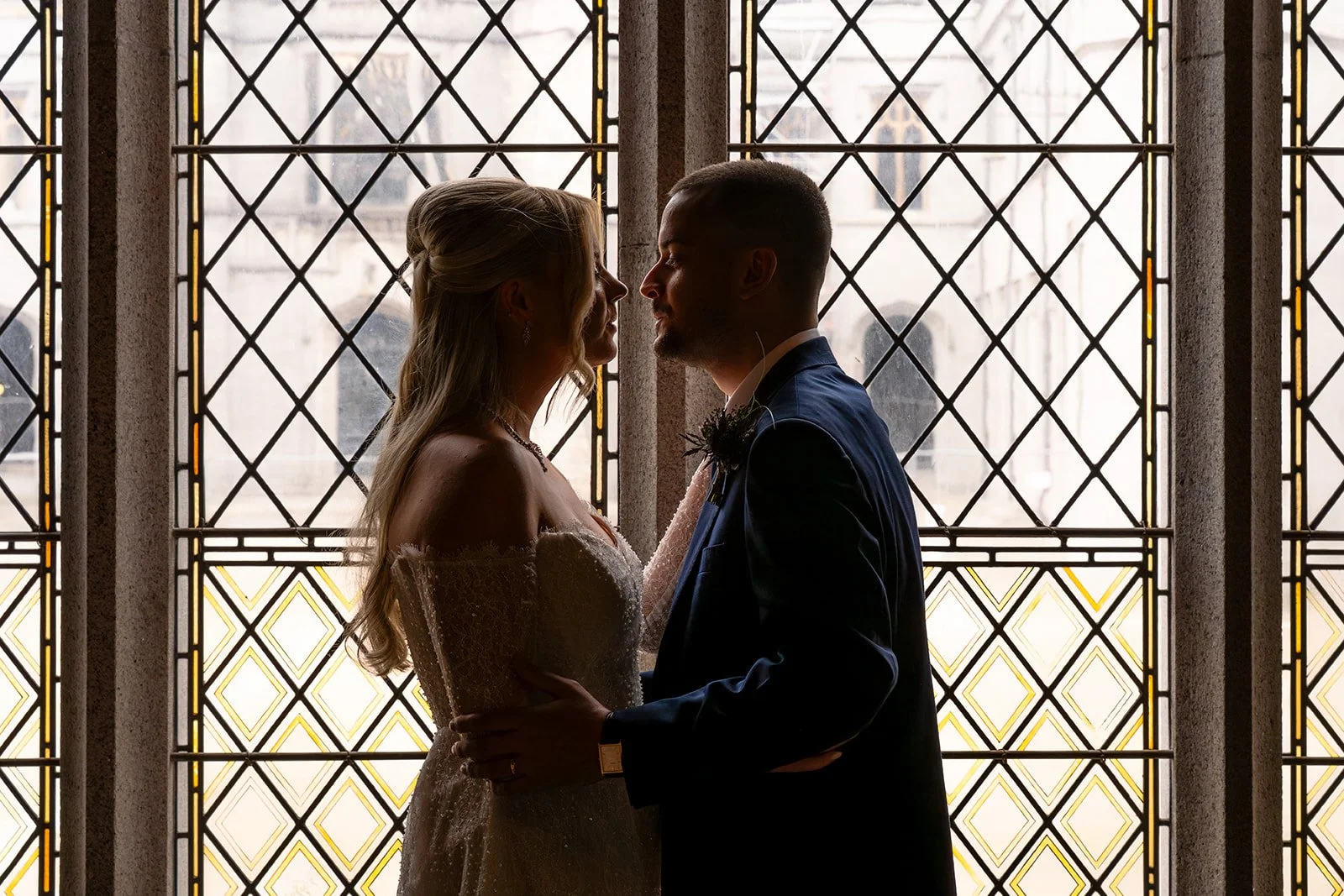 A couple in wedding attire standing close together in front of a decorative stained glass window, facing each other and holding each other