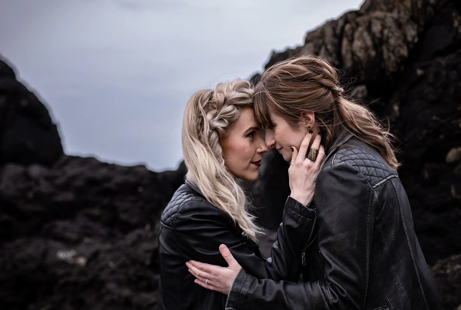 Two women with their foreheads and noses touching, gazing into each other's eyes, wearing black leather jackets, with dark rocky landscape in the background.