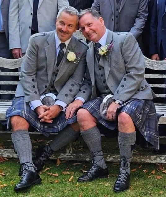 Two men dressed in gray kilts, gray jackets, and gray vests sitting on a wooden bench, smiling and leaning towards each other, with a group of men in similar attire standing behind them. They are outdoors on grass with fallen leaves.