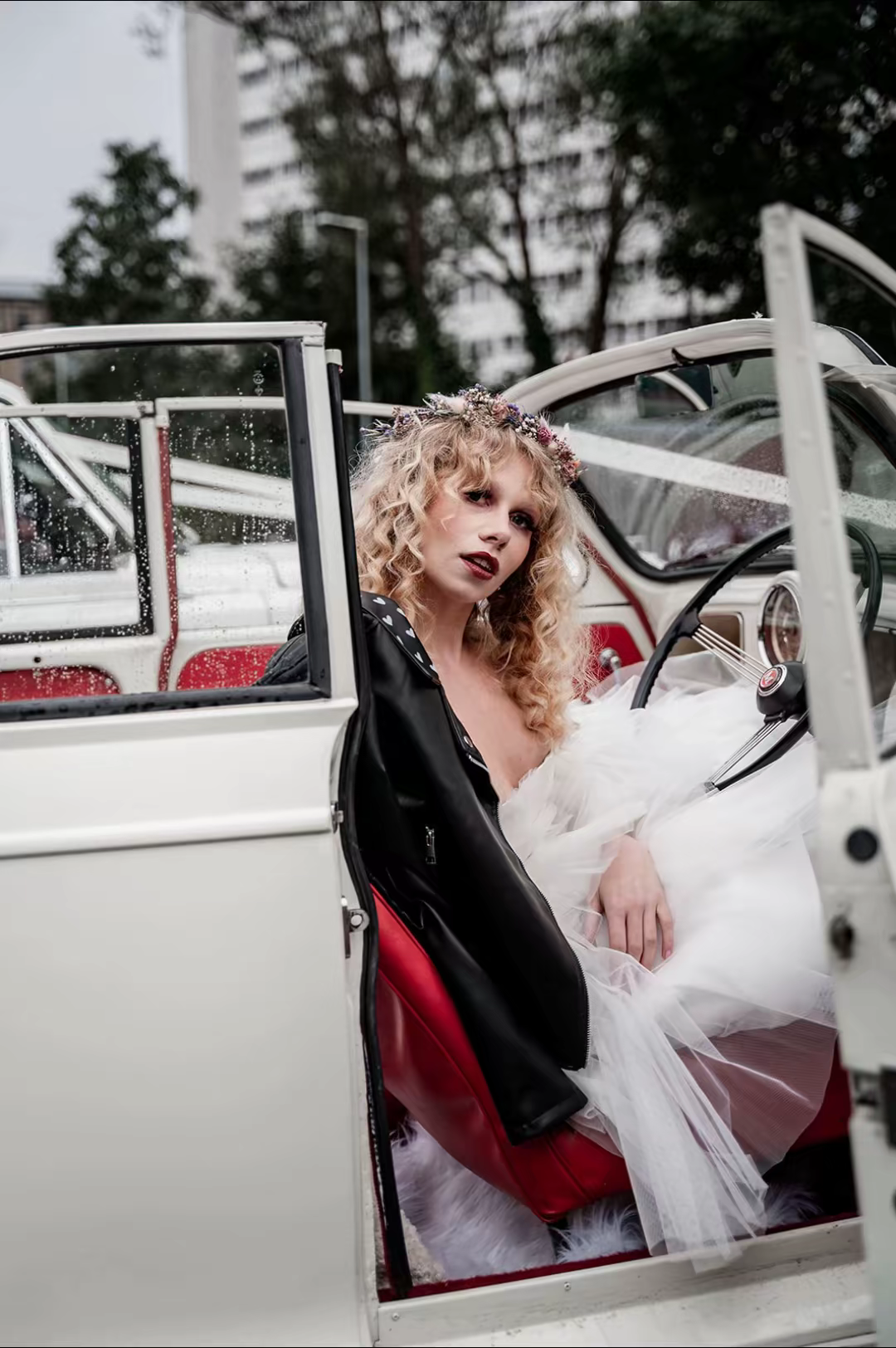 A woman with curly blonde hair sits in the open door of a white vintage car, wearing a white dress and a floral crown, with a black leather jacket draped over her shoulder, in an urban setting with tall buildings and trees in the background.