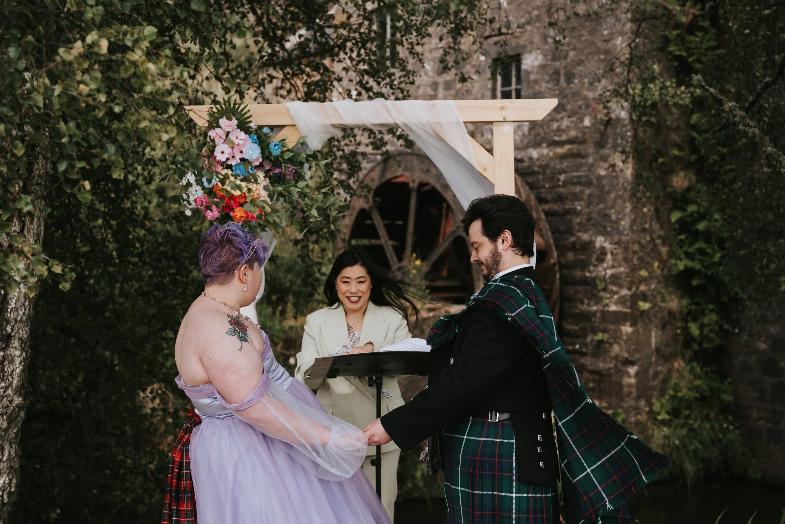 A couple getting married outdoors, holding hands and facing each other, with a woman officiant behind them, smiling. The ceremony is under a wooden arch decorated with colorful flowers.