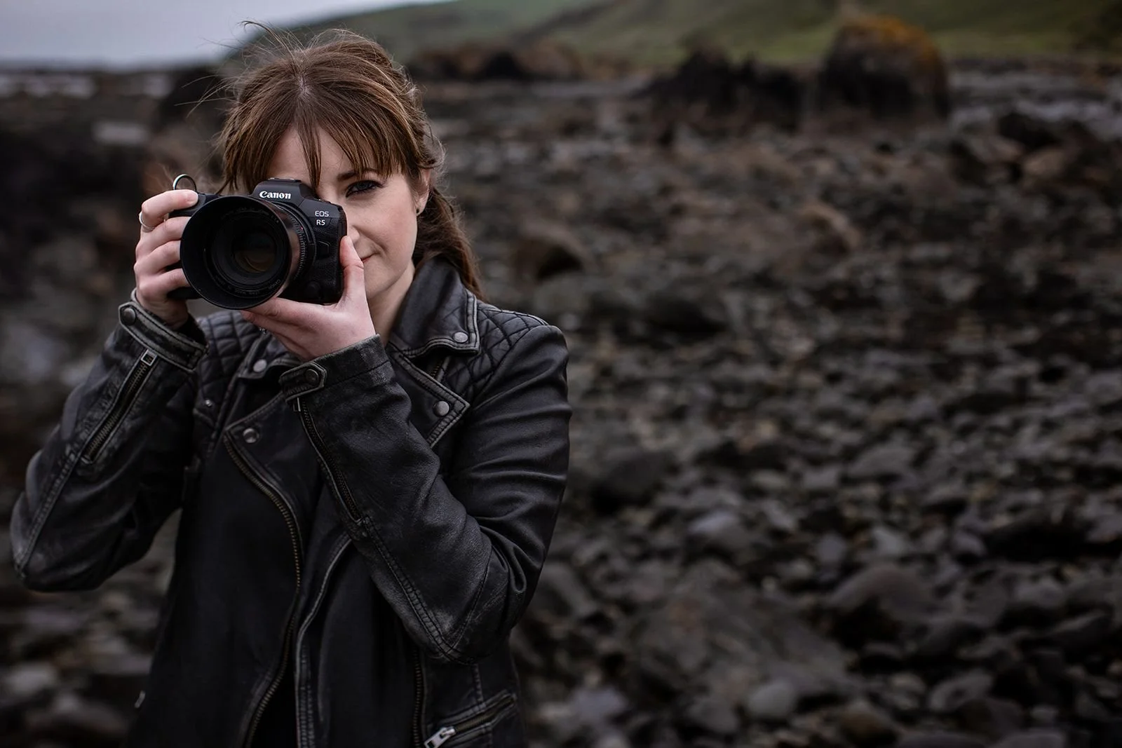 A woman with brown hair and a black leather jacket holding a Canon camera up to her face, standing outdoors on rocky terrain with a blurred background of rocks and greenery.