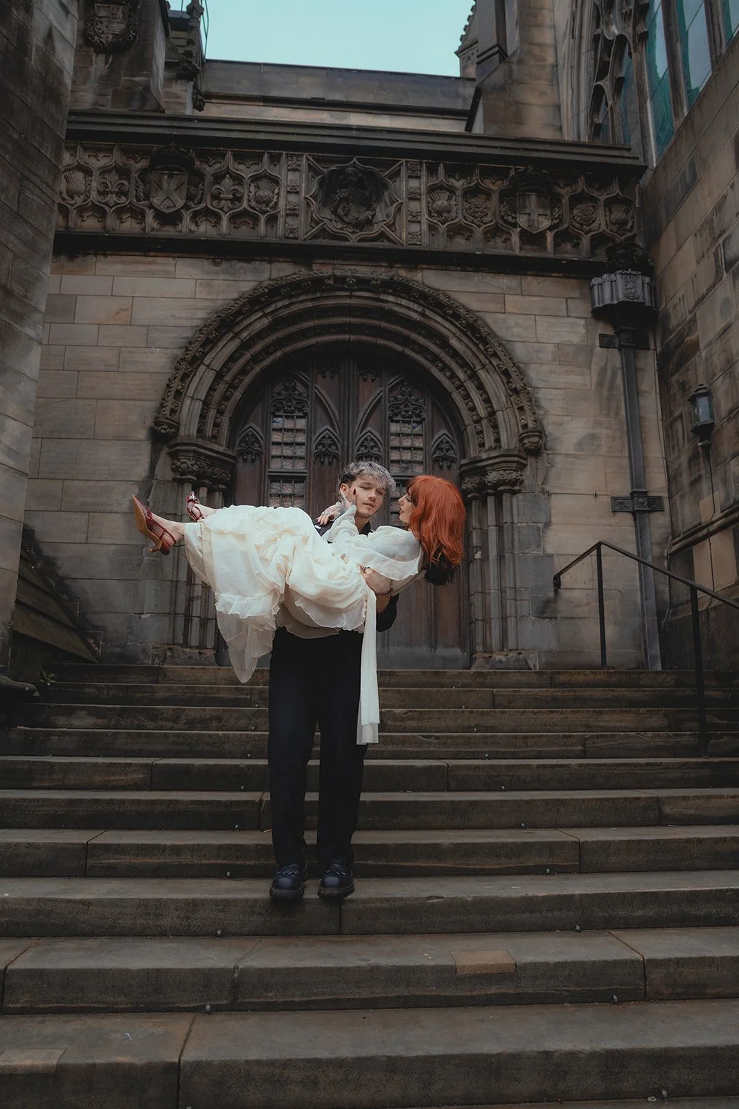 A man in black clothing holding a woman in a white dress in front of a historic stone building with a large arched doorway and intricate carvings.