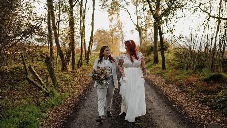 Two women, one in a wedding dress and the other holding a bouquet, walking hand-in-hand on a dirt path in a wooded area during sunset.