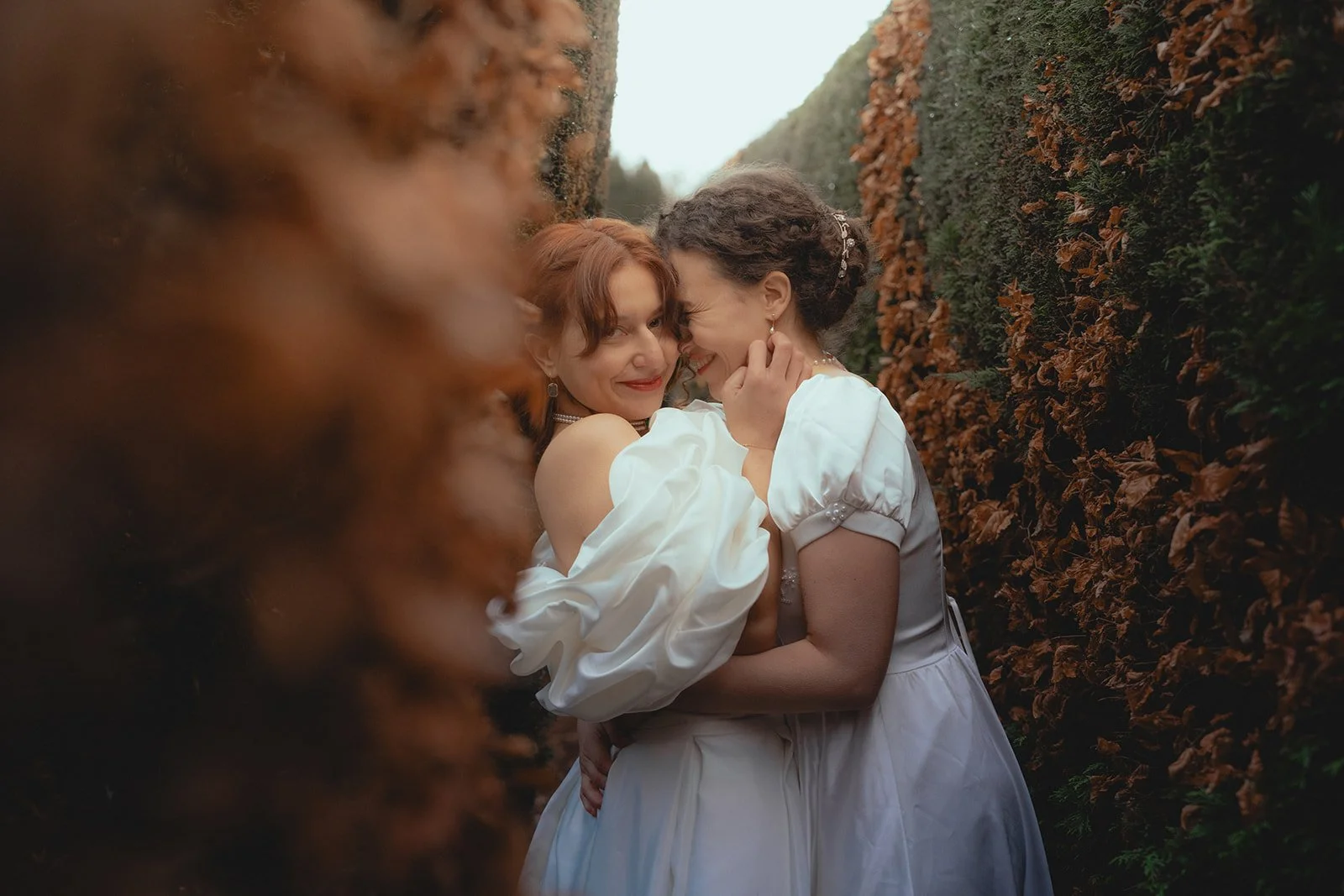 Two women dressed in white embrace affectionately between two tall hedges, smiling and leaning their foreheads together.