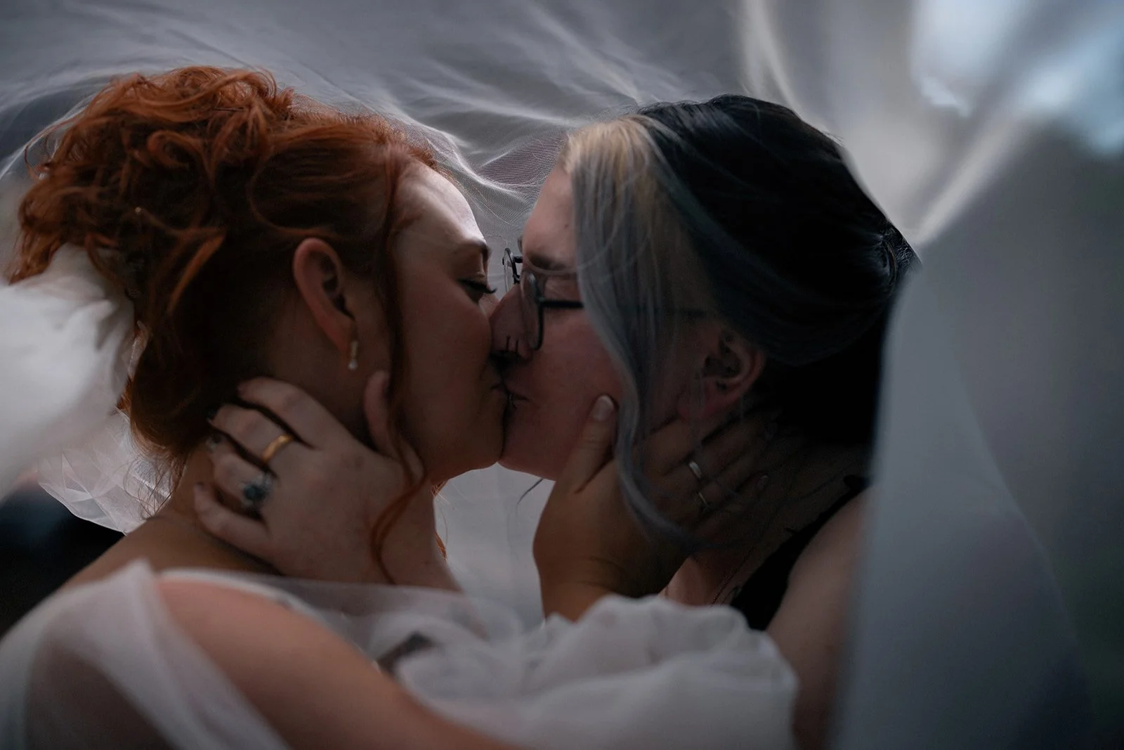 A couple sharing a kiss, with the woman having red hair and the man wearing glasses, both embracing in an intimate moment indoors.