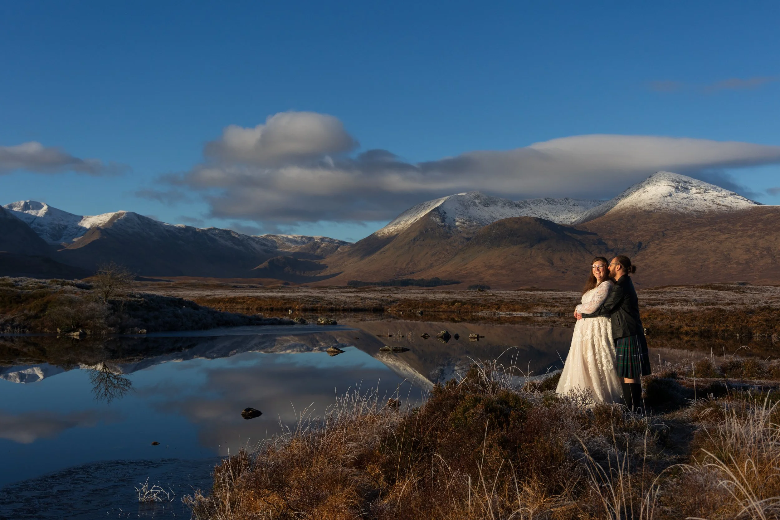 A couple in wedding attire standing by a reflective body of water with snow-capped mountains and a blue sky with clouds in the background.