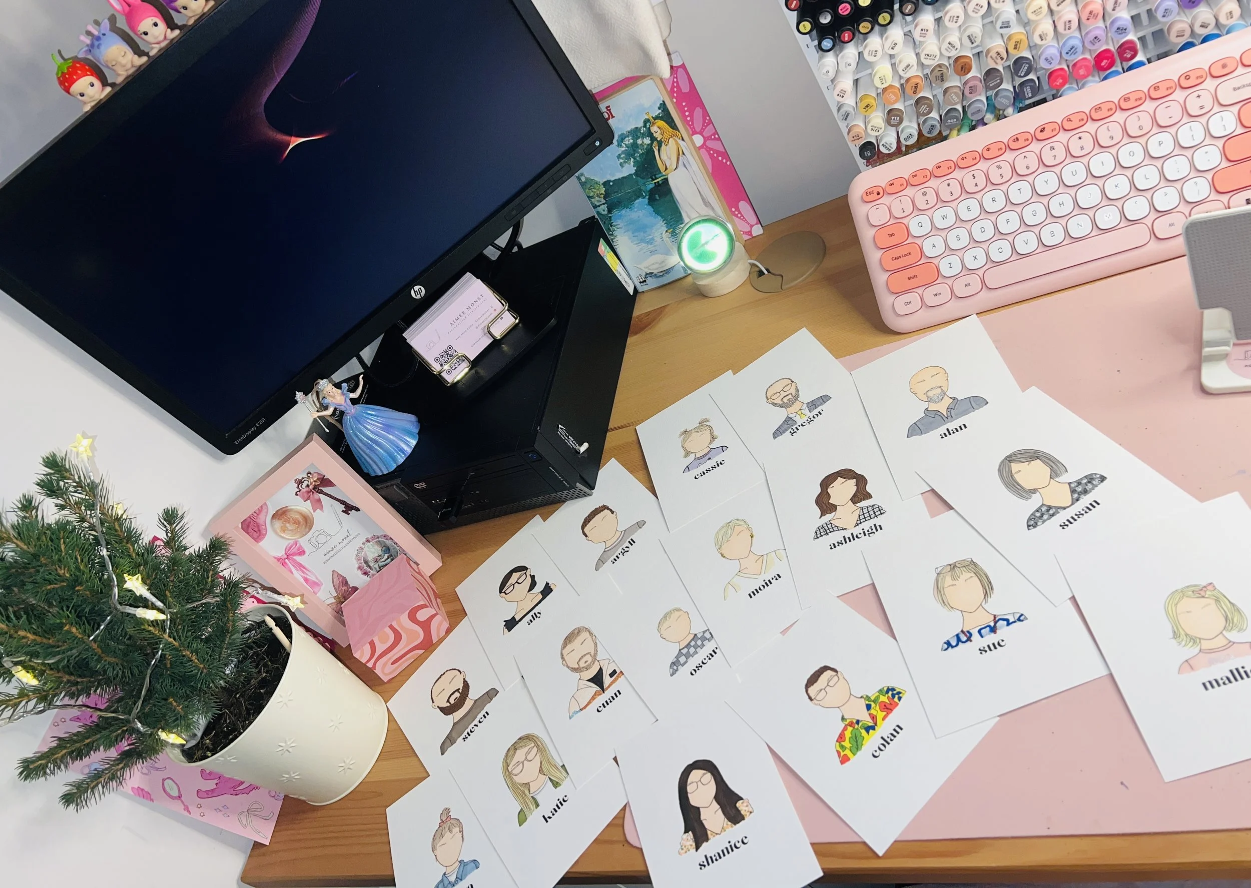 A workspace with a computer monitor, a small Christmas tree, various illustrated name tags, a pink keyboard, a color pen holder, a small figurine, and colorful decorations on the desk.