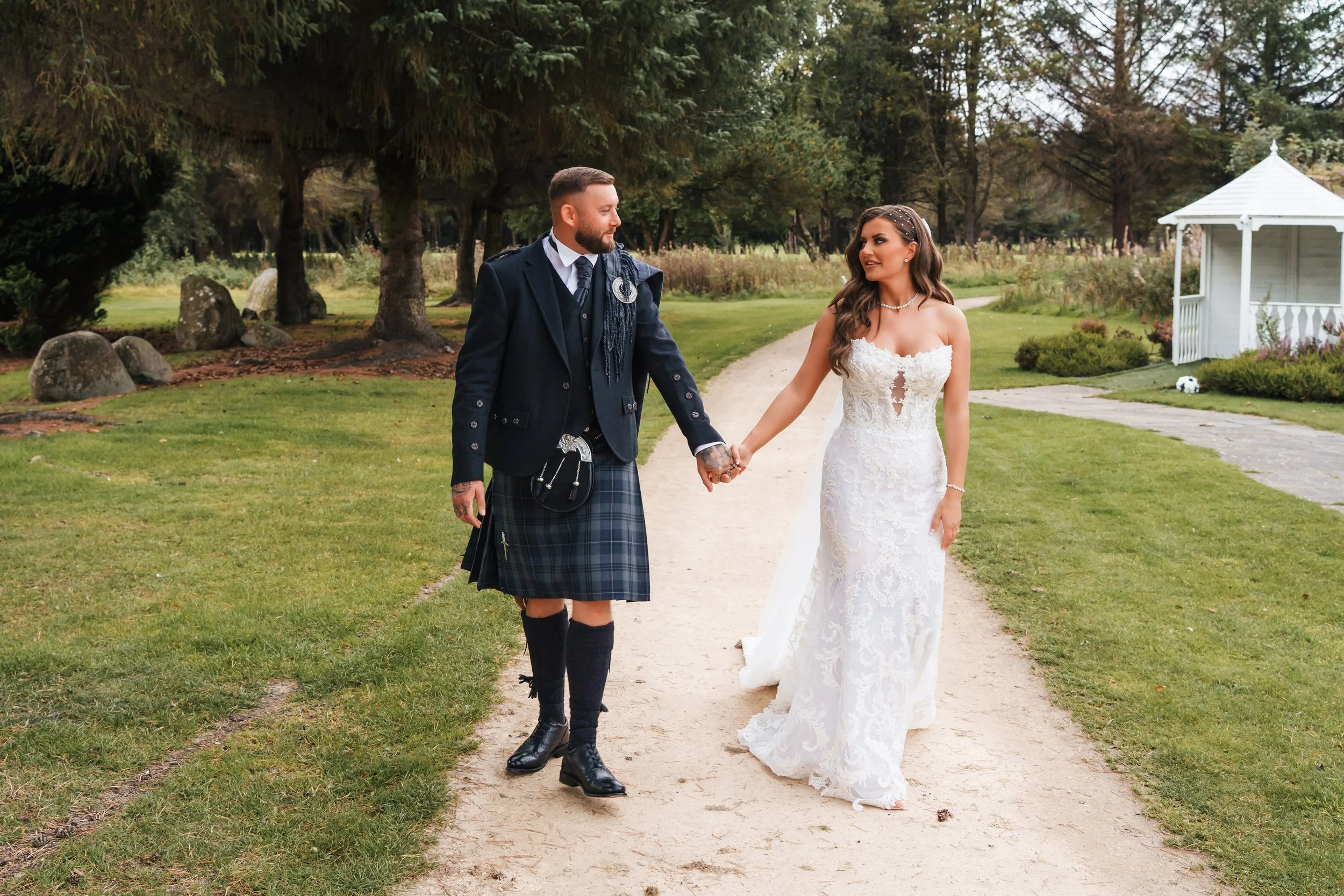 A couple walking hand in hand outdoors on a dirt path, with a woman in a white wedding dress and a man in a traditional Scottish kilt and jacket.