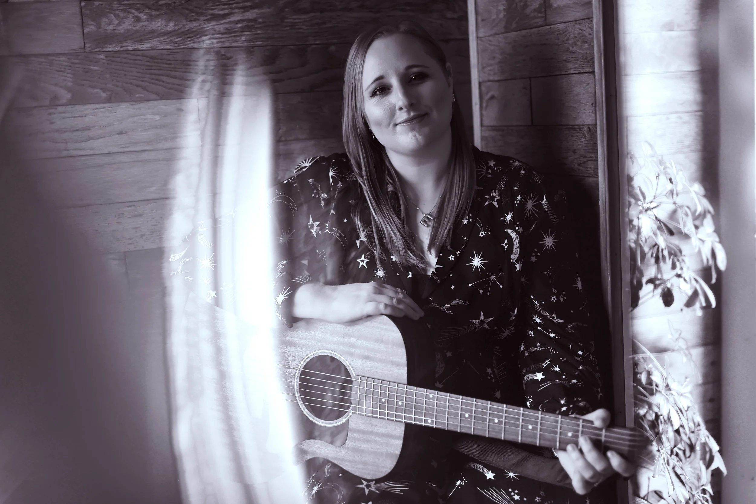 A woman with long hair sitting against a wooden wall, holding an acoustic guitar and smiling softly at the camera, with a potted plant nearby.