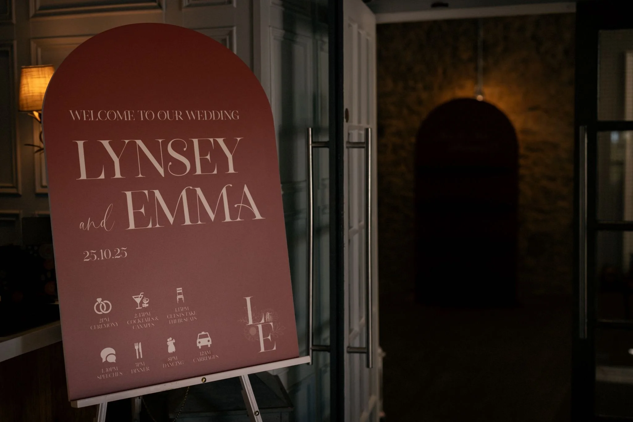 Wedding welcome sign with names Lynsey and Emma, date October 25, 2025, in a dimly lit venue entrance, with icons indicating ceremony, drinks, meals, dancing, and parking.