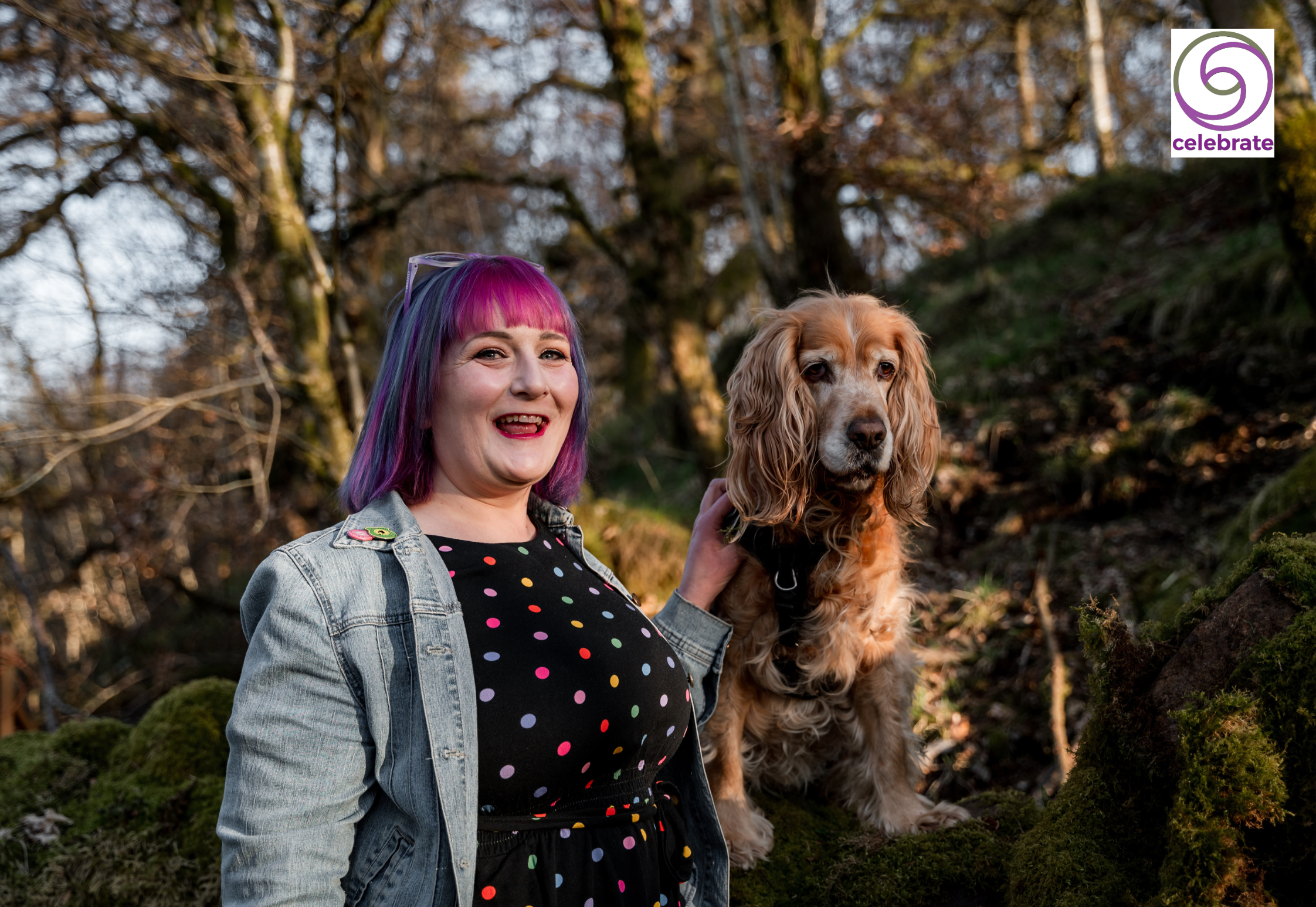 A woman with pink and purple hair smiling outdoors next to a brown and white Cocker Spaniel dog, with trees in the background.