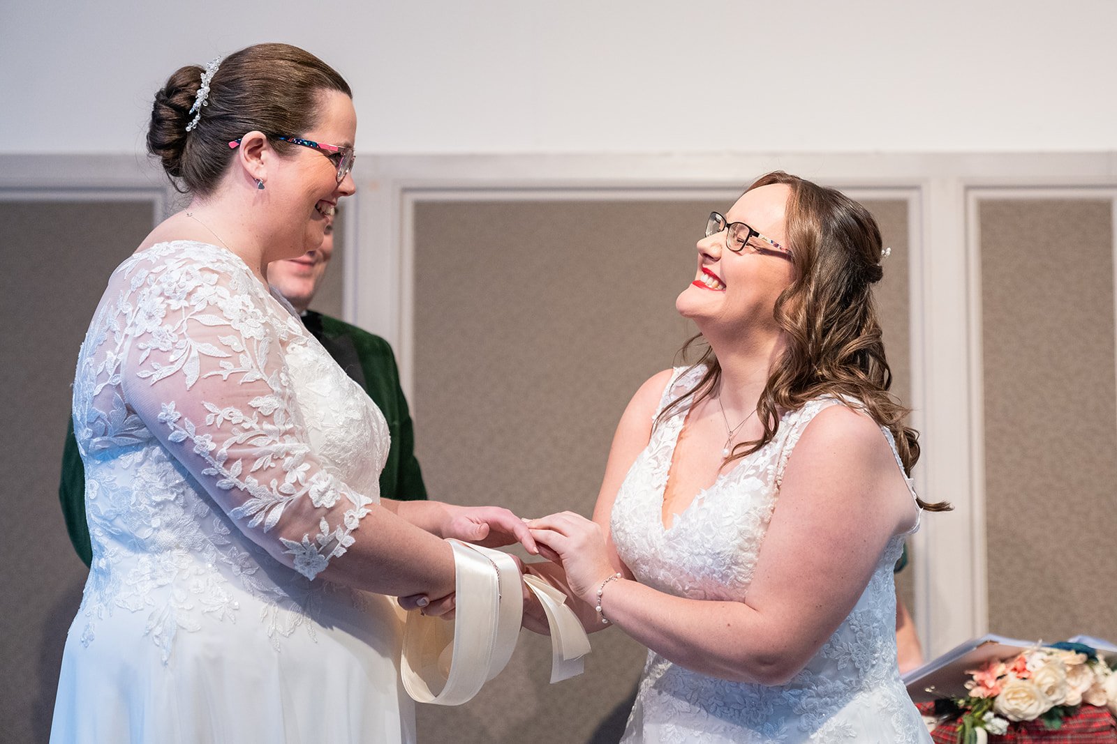 Two women in wedding dresses hold hands and smile at each other during a wedding ceremony, with a man partially visible in the background and a table of flowers nearby.