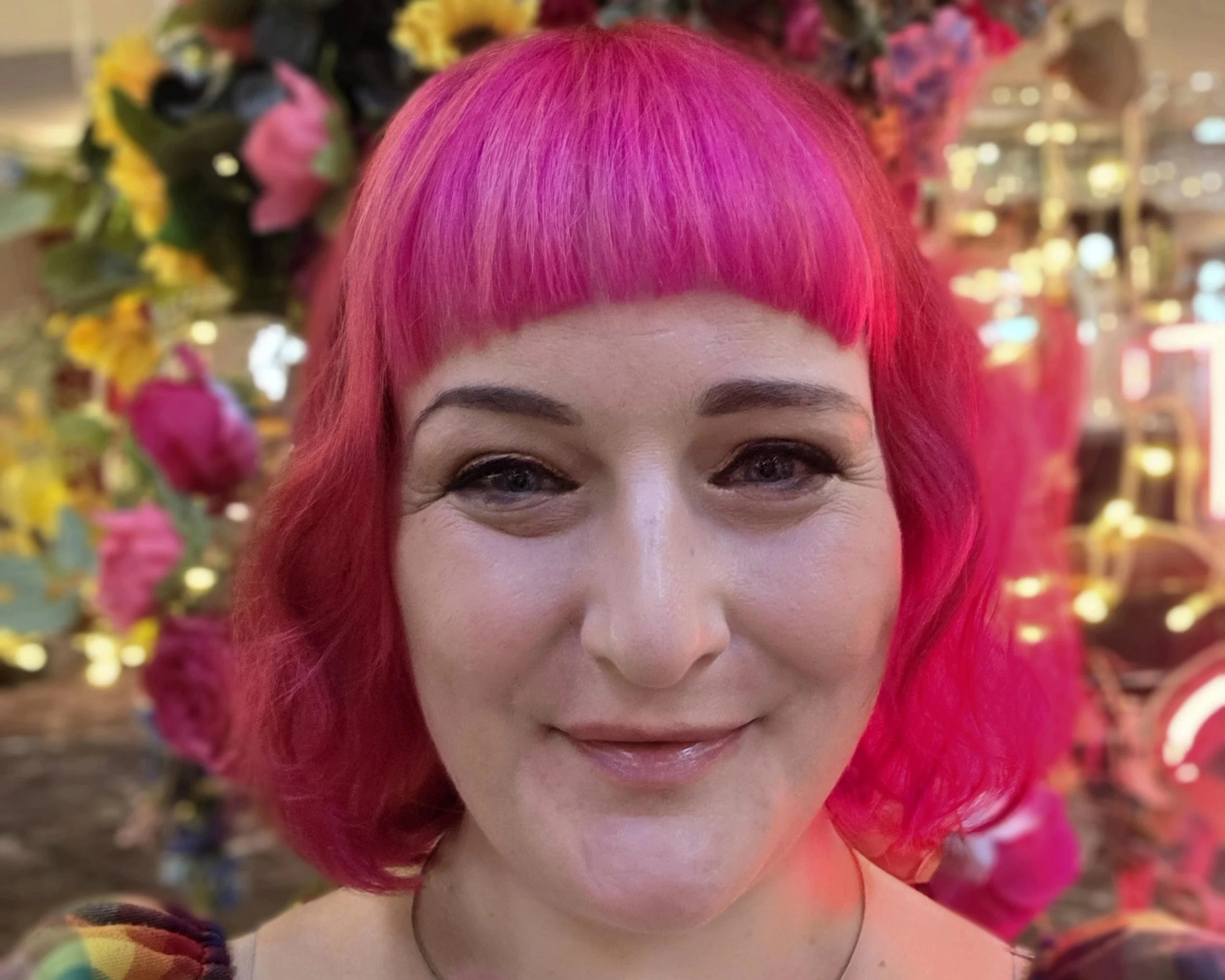 A woman with bright pink hair styled in a bob with bangs, smiling in front of a colorful floral display in an indoor setting.