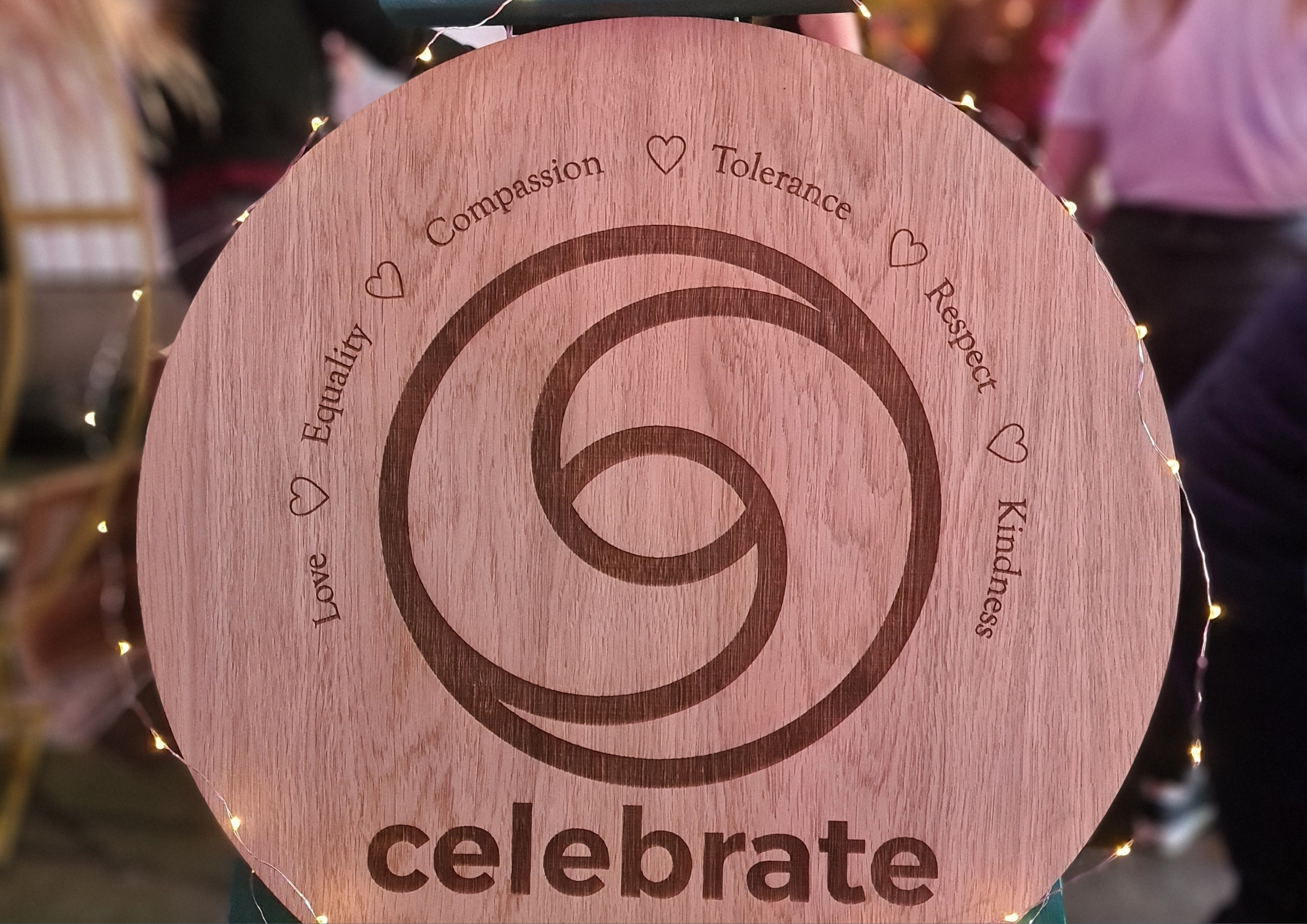 Decorative round wooden sign with the word 'celebrate' at the bottom and concentric circles in the center, surrounded by words 'Love,' 'Equality,' 'Compassion,' 'Tolerance,' 'Respect,' 'Kindness,' and small heart icons, with string lights around the edge.