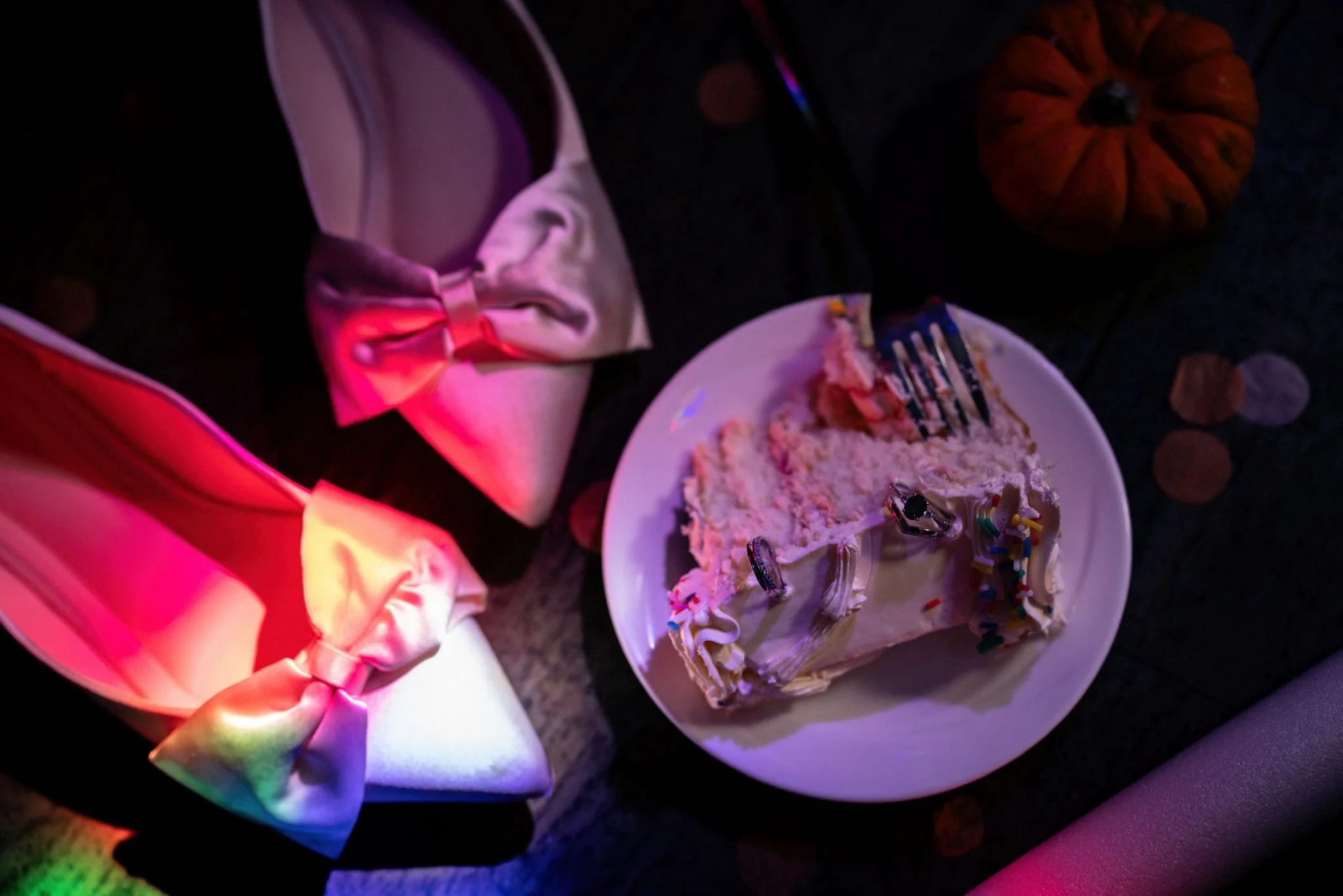 A slice of birthday cake with white frosting and colorful sprinkles on a white plate, surrounded by party decorations including a pumpkin and wrapped gifts with ribbons, all under pink and purple lighting.