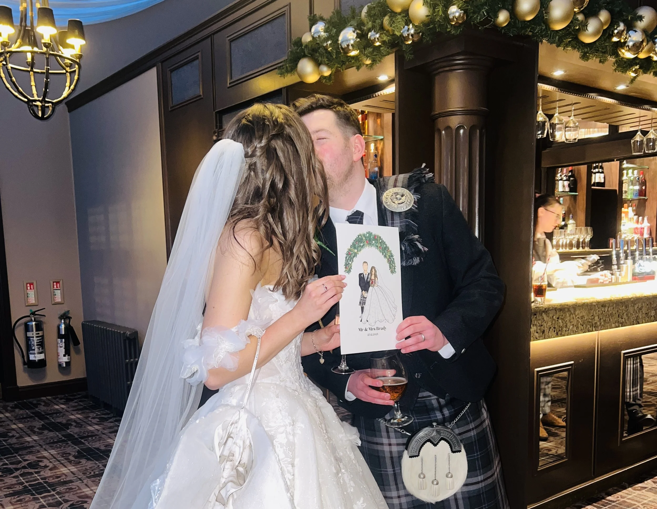 A bride and groom sharing a kiss at their wedding reception, holding a wedding card with a groom and bride illustration and white Christmas wreath, with a bar in the background decorated with festive ornaments.