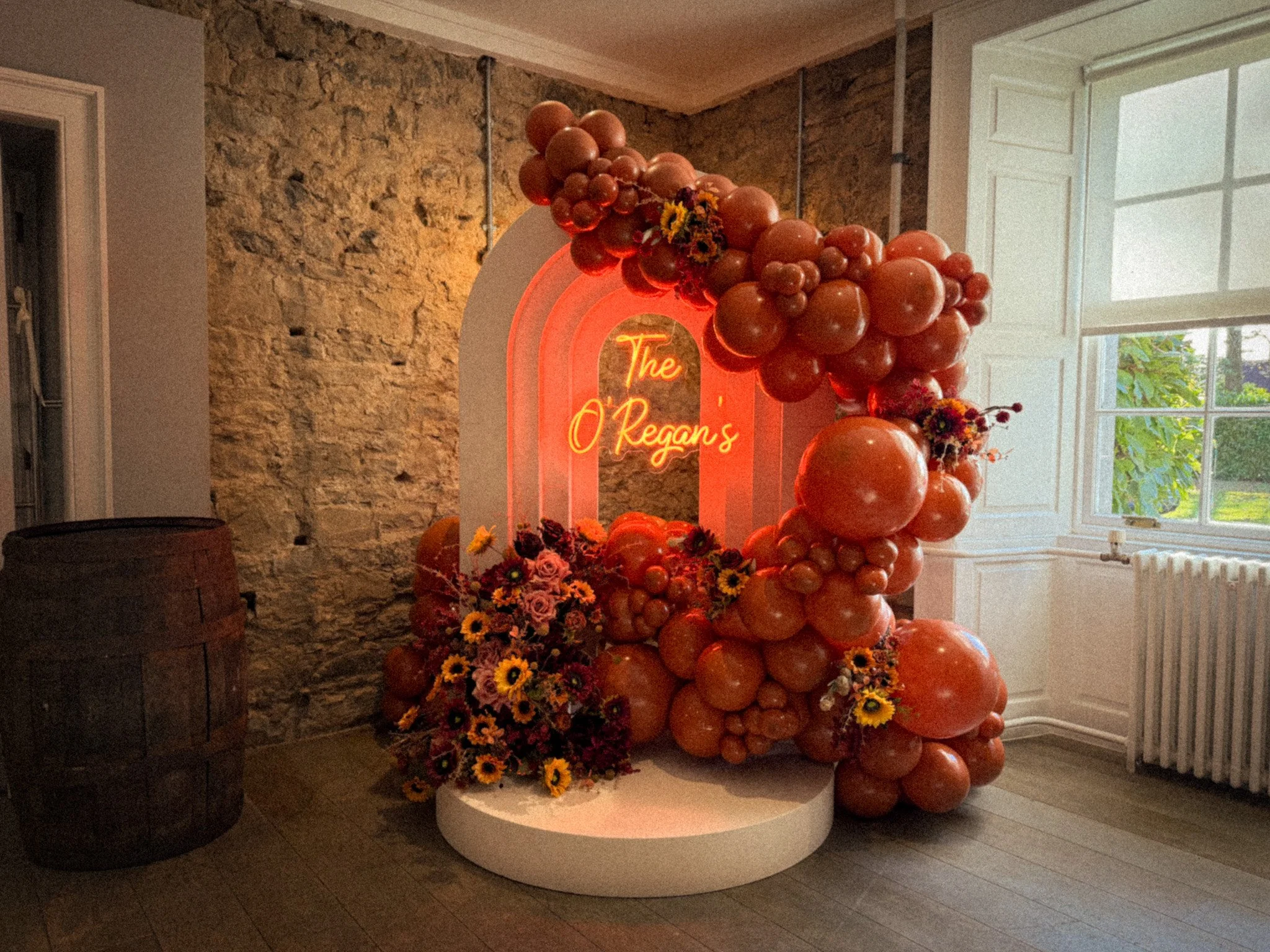 Decorative display with orange balloons, flowers, and a neon sign reading 'The O'Regan's' inside a room with stone wall and large window.