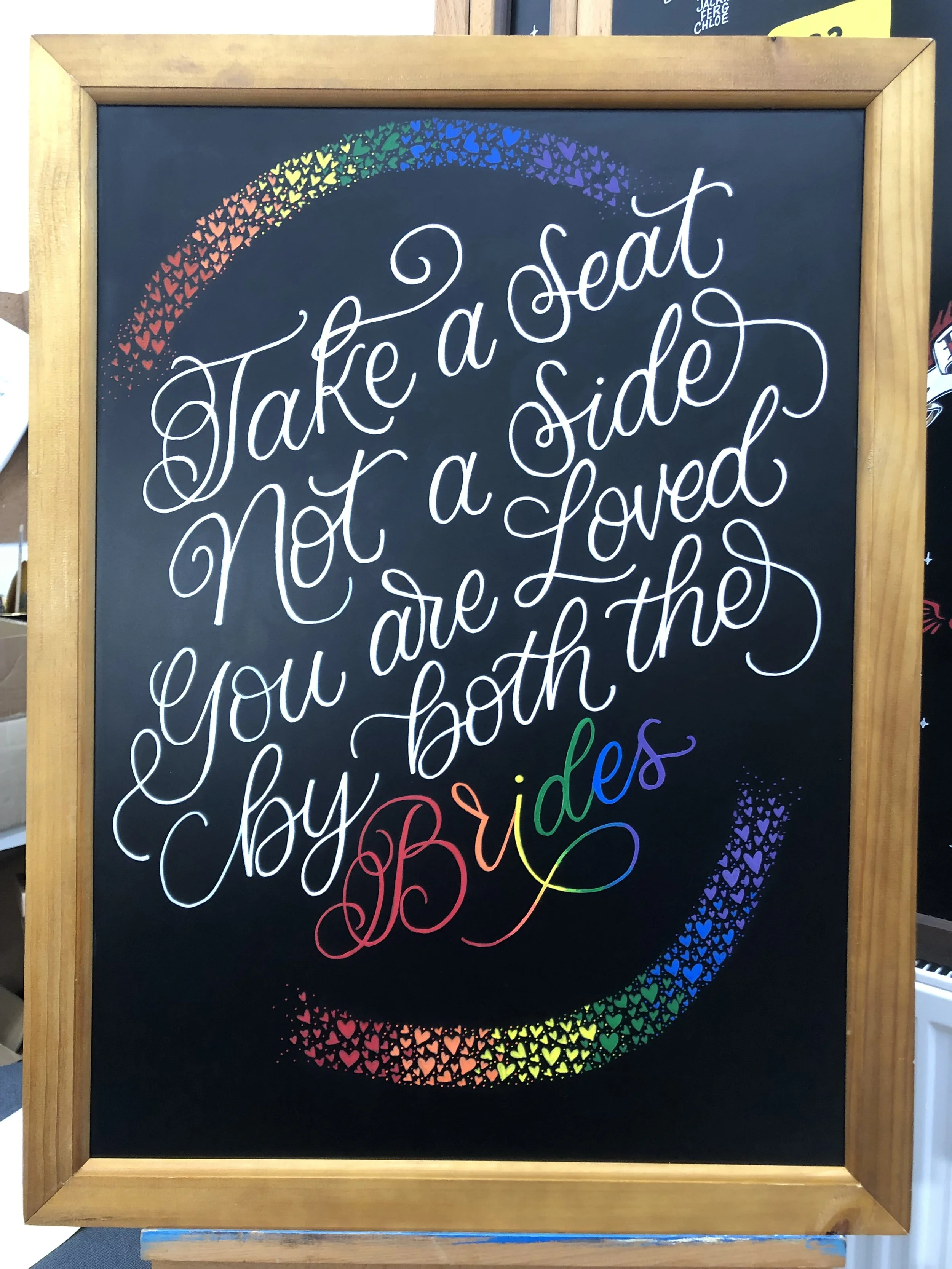 Colorful chalkboard sign with a handwritten message: "Take a seat Not a Side You are Loved by both the Brides" surrounded by a rainbow made of tiny hearts.