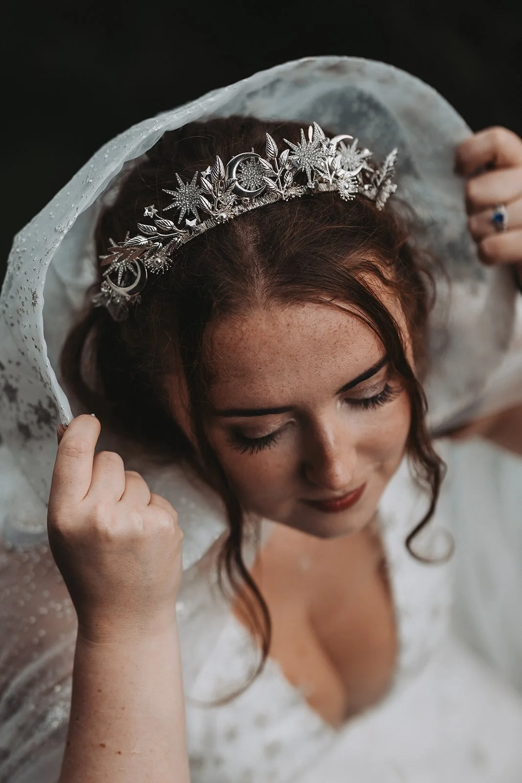 Close-up of a bride with a silver tiara, holding her veil, with her eyes closed and makeup on her face.