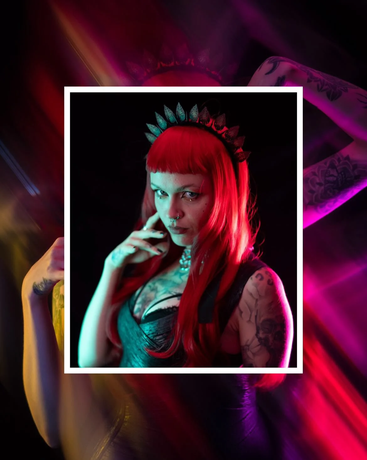 A woman with bright red hair, wearing a crown-like headband, looking at the camera with a confident expression, in a dark setting with colorful neon lighting.