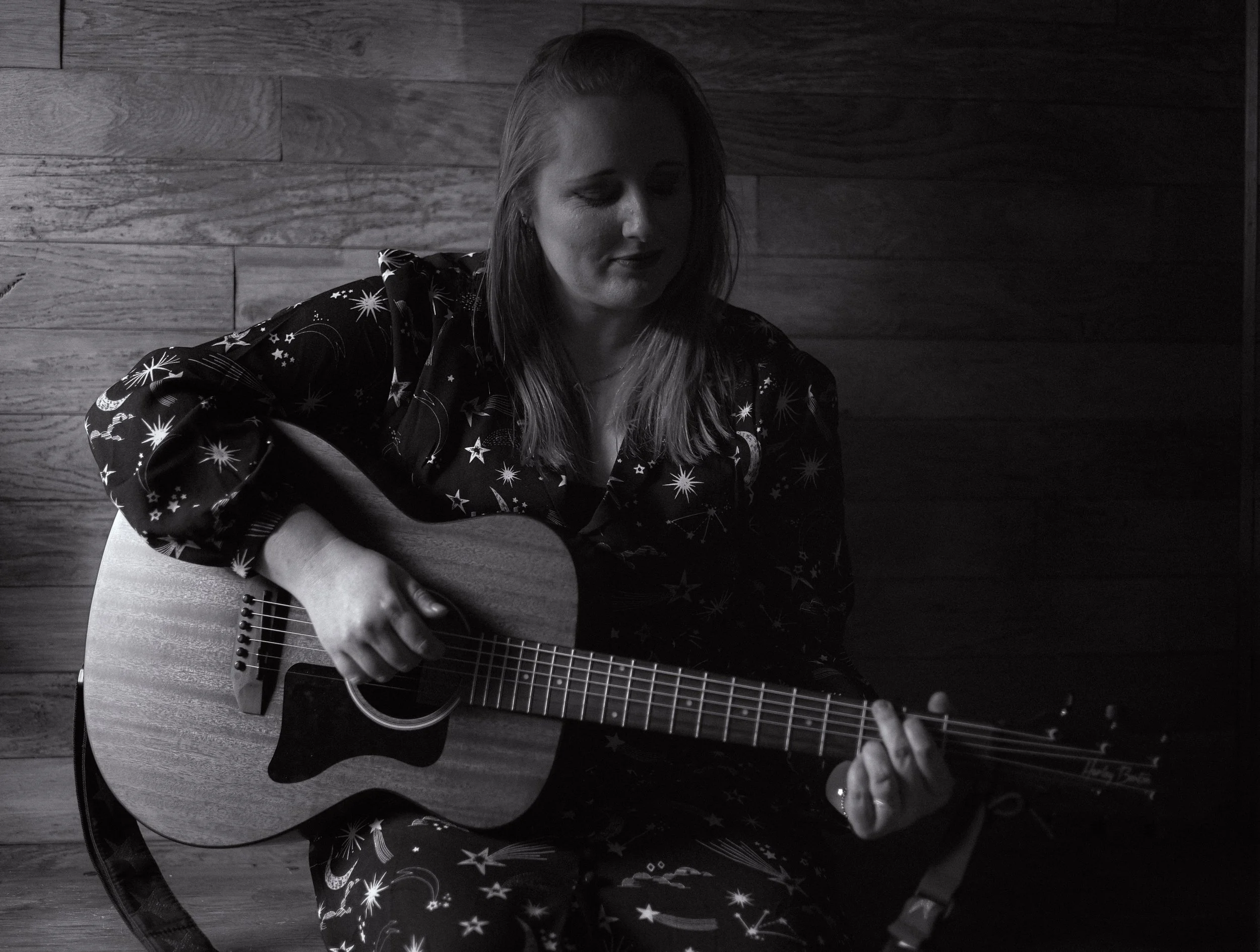 A woman playing an acoustic guitar while sitting in front of a wooden wall, wearing a dark pajama with star and moon patterns.