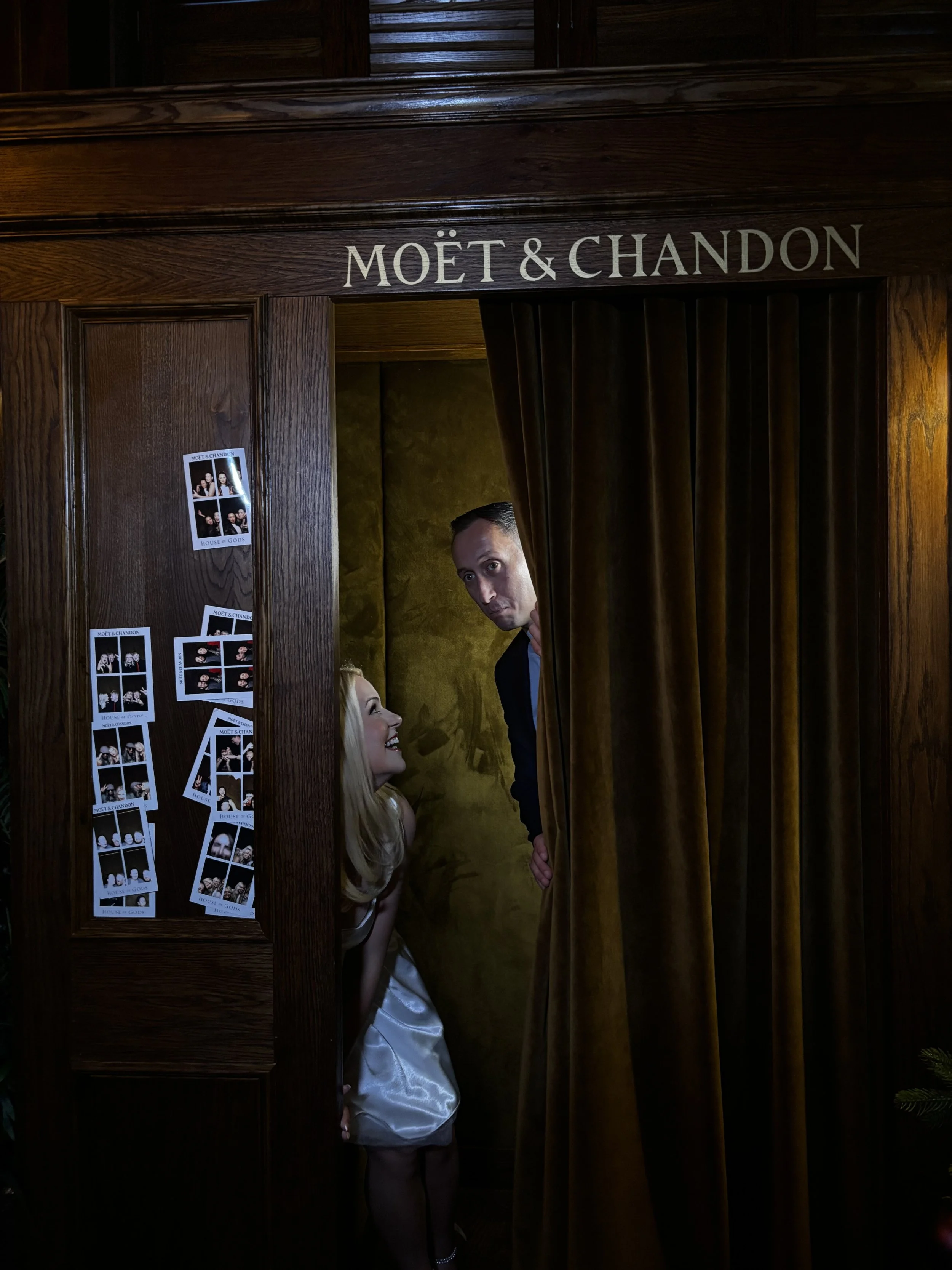 Two people peeking out from behind a curtain in a Moët & Chandon event setup, with photo booth strips on the wall.