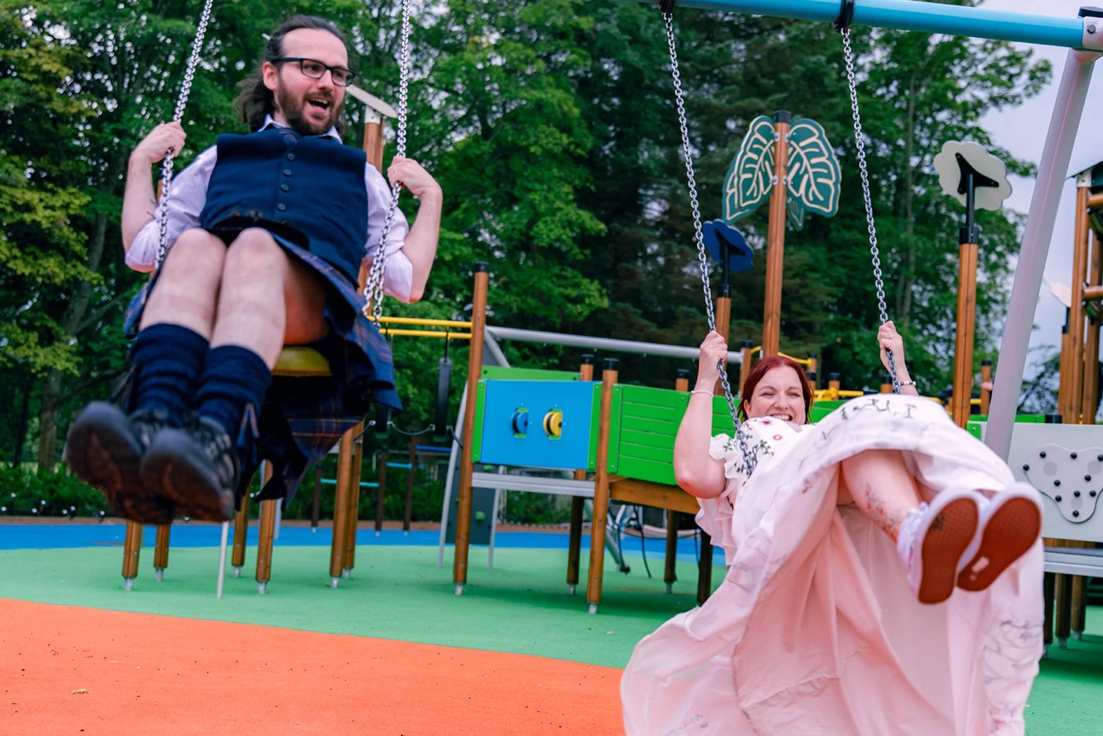 Two people, a man and a woman, are swinging on a playground swing set. The man is dressed in a traditional Scottish kilt and vest, smiling and holding the chains. The woman is wearing a pink floral dress, smiling widely, and holding the chains while swinging high. The playground has colorful flooring and background is filled with green trees.