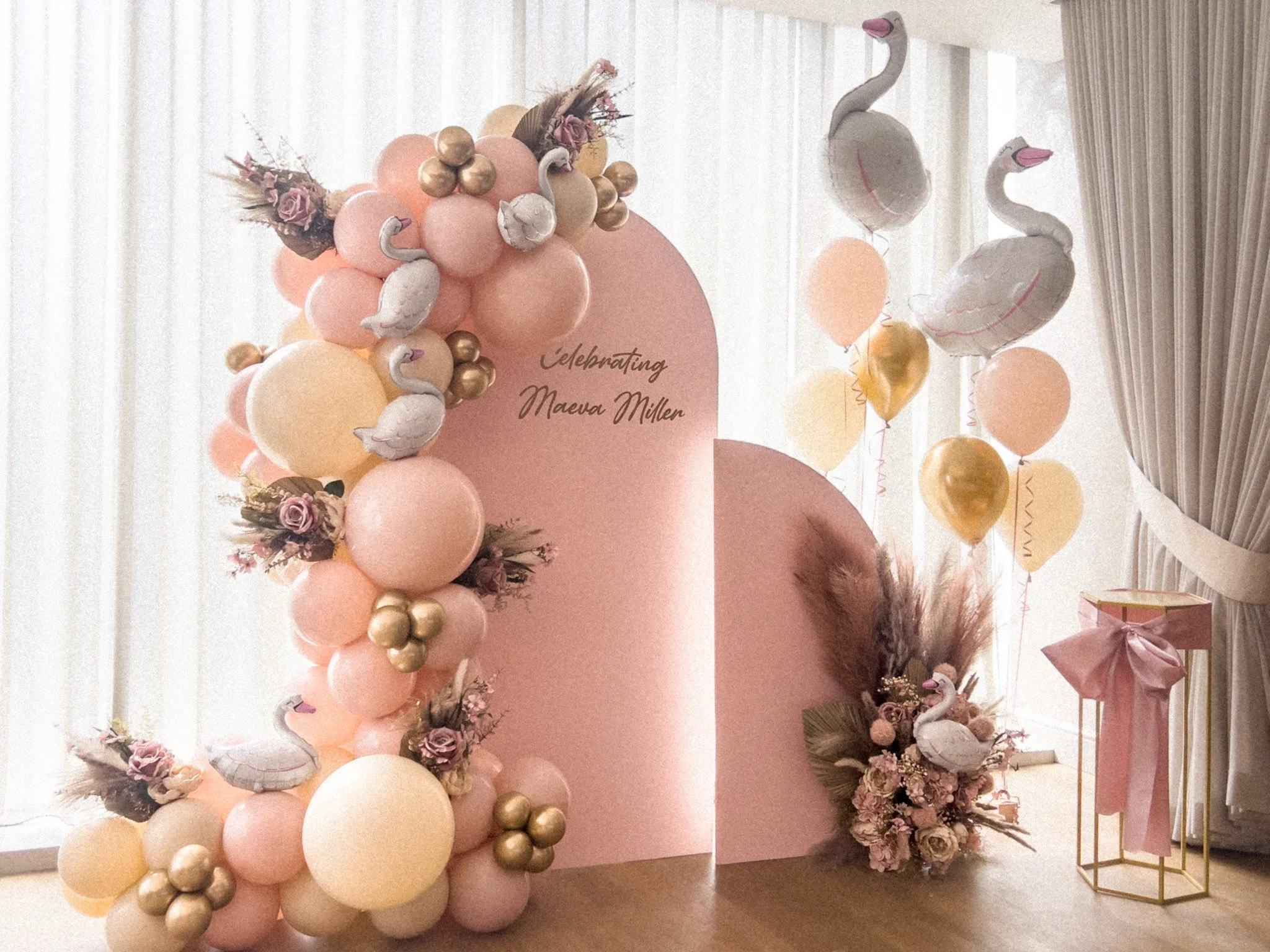 Decorative setup with pink and gold balloons, swan and floral arrangements, and a pink backdrop with the text 'Celebrating Maeva Miller' for a celebration or party.