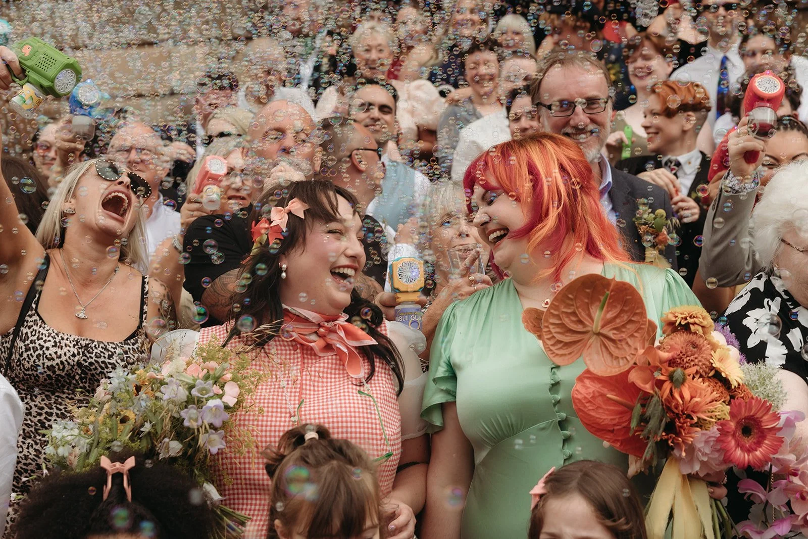 Group of people celebrating outdoors with bubbles floating around, some holding colorful bouquets of flowers, smiling and laughing.