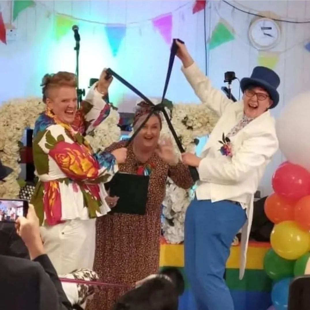 Three people celebrating at a festive event, one woman holding a microphone, one woman in colorful clothing, and a man in a white blazer and top hat. They are smiling and appear to be in a joyful mood, with colorful balloons and decorations in the background.