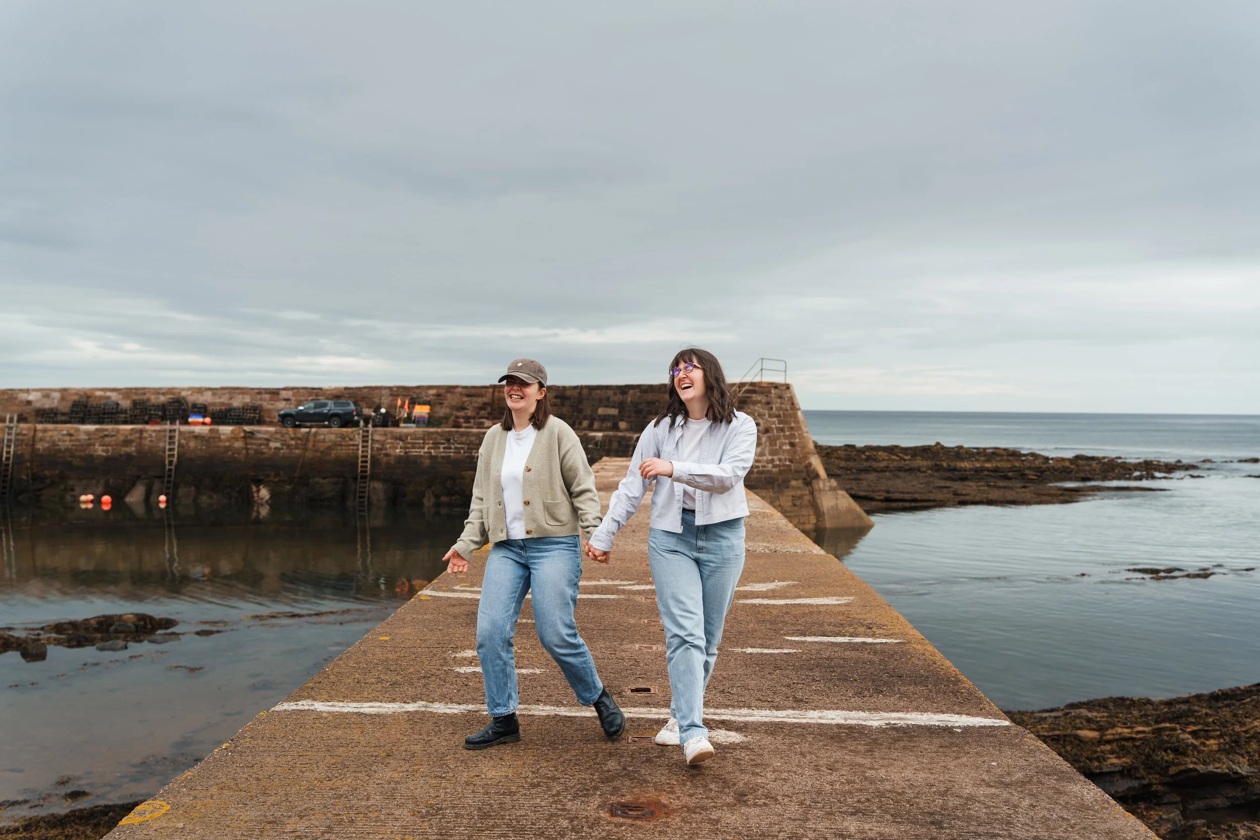 Two women holding hands and smiling while walking on a concrete pier by the water on an overcast day.