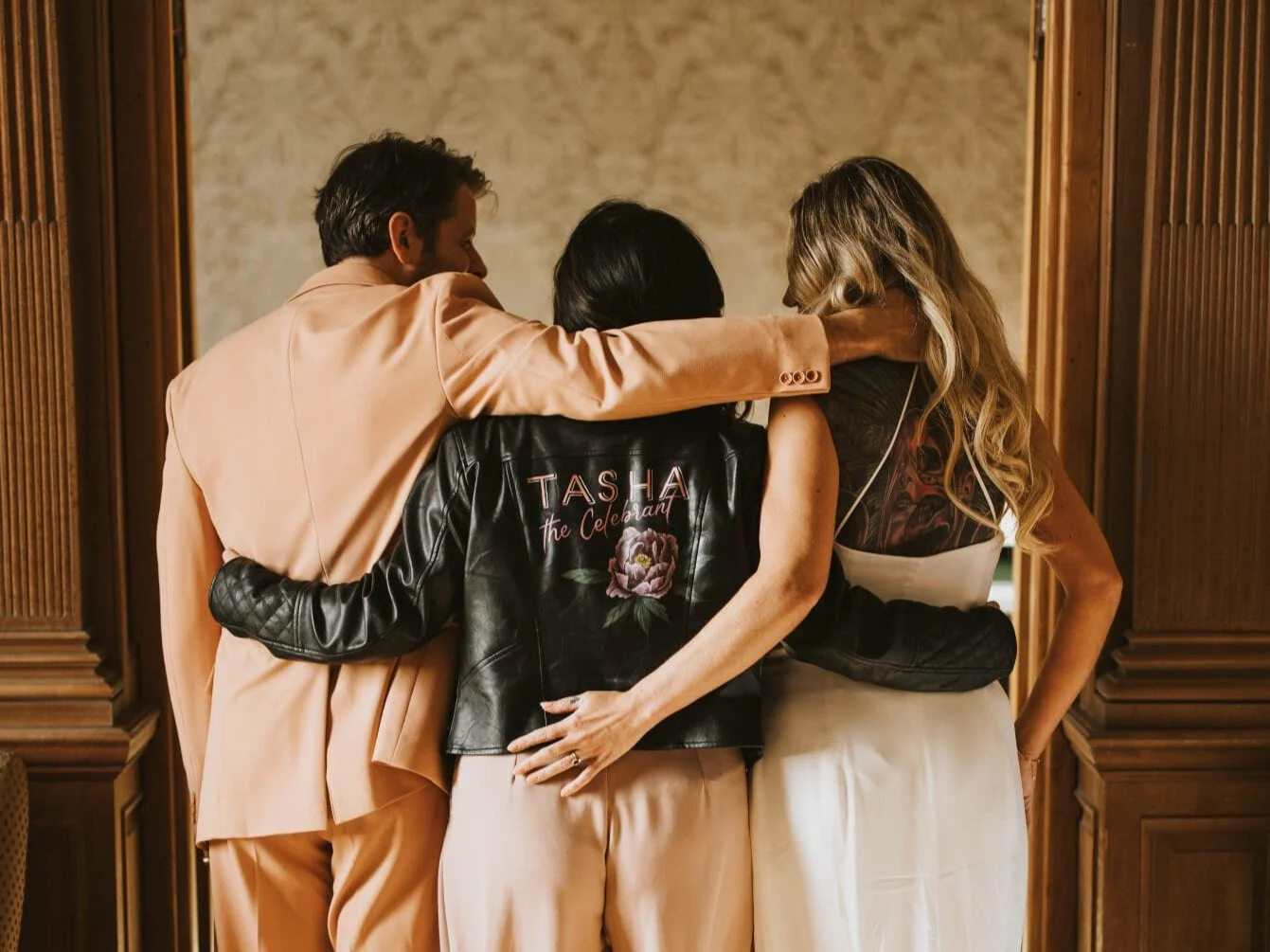 Three people embracing in a hugging circle with arms around each other, standing indoors in front of wooden paneling.