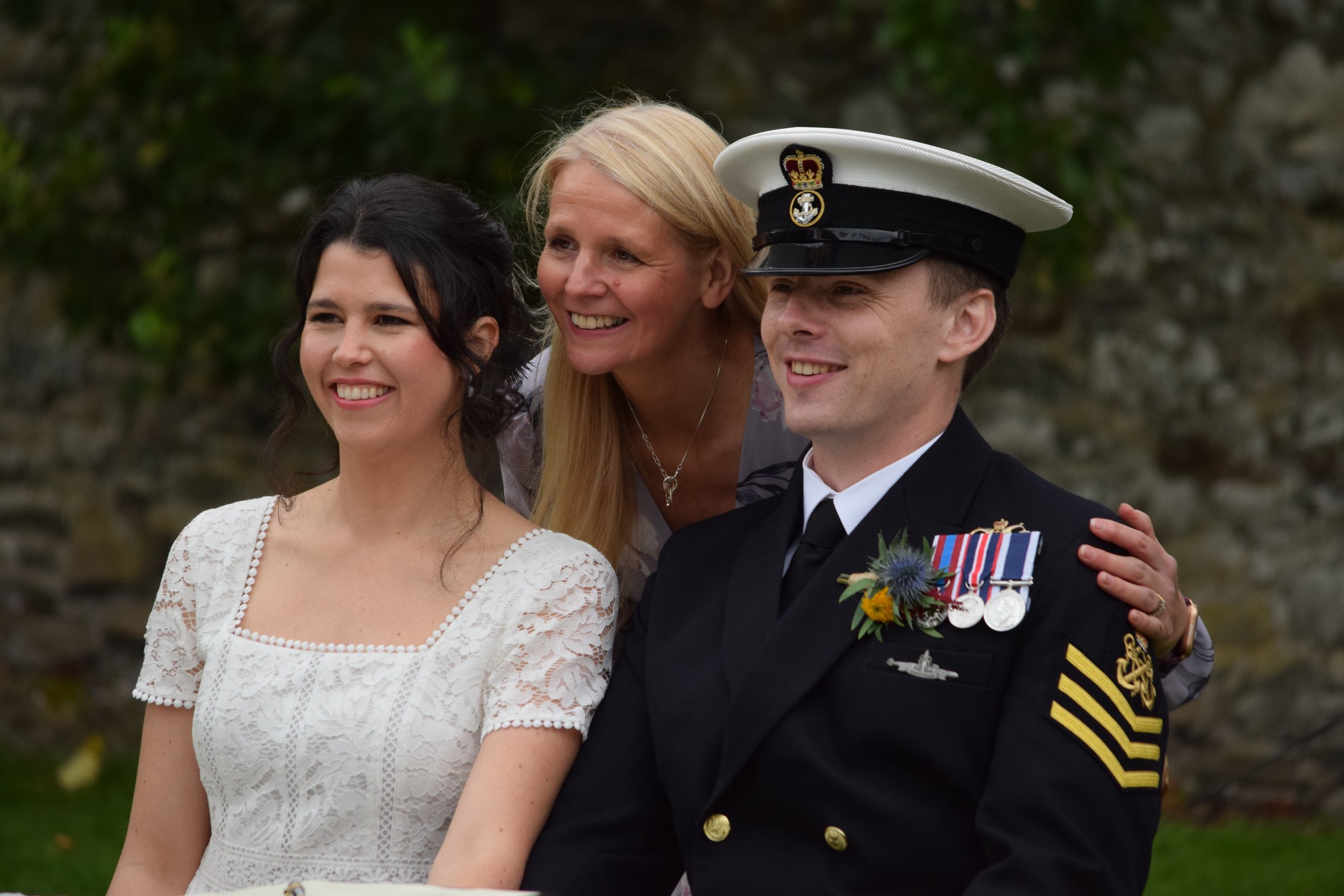 Three people smiling at a gathering, with a woman in a white dress, another woman with blonde hair, and a man in a Navy uniform wearing medals and a white hat with a black band, outdoors near a stone wall.
